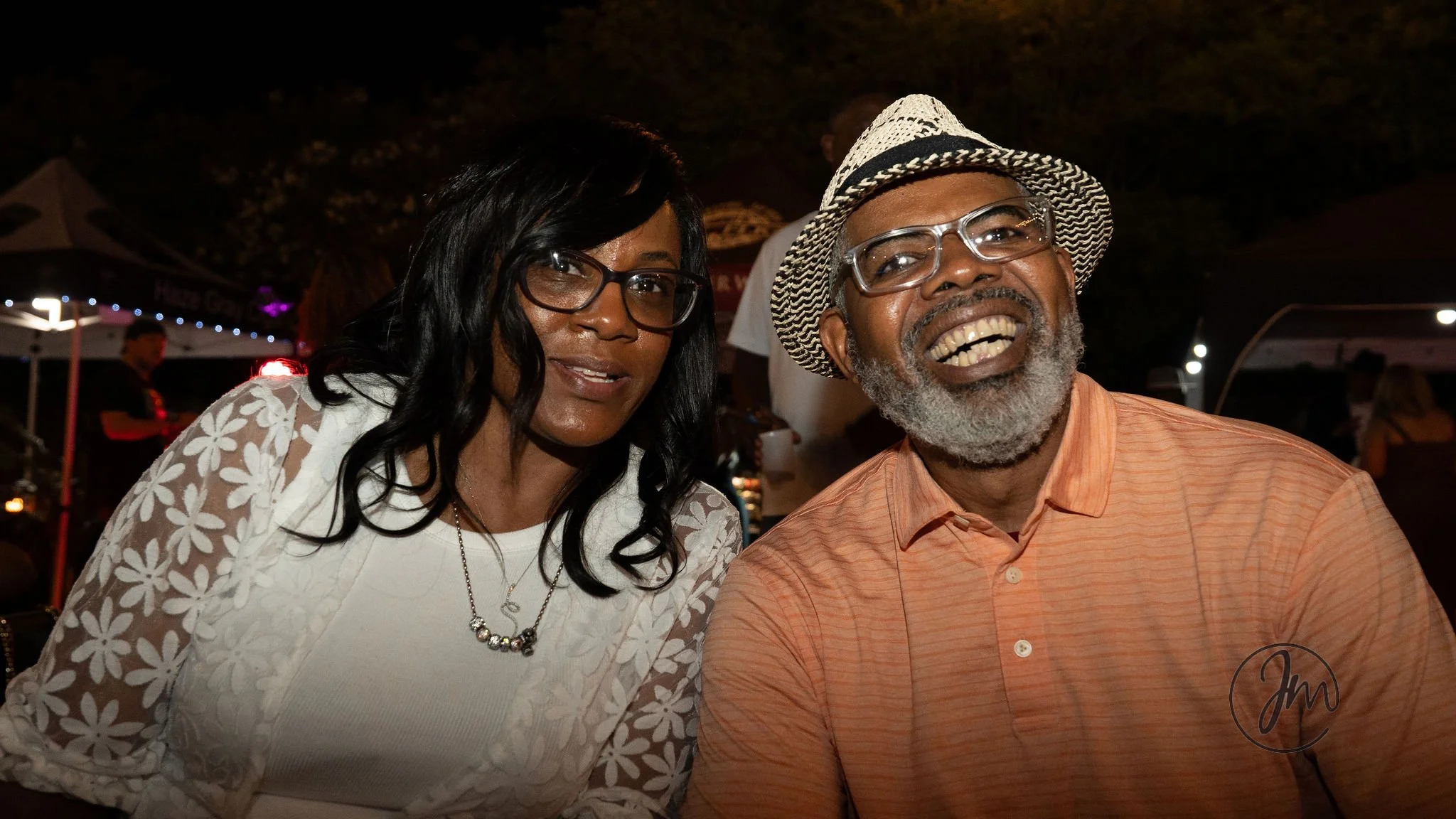 Two people smiling at an event at night, outdoor setting with tents and string lights.