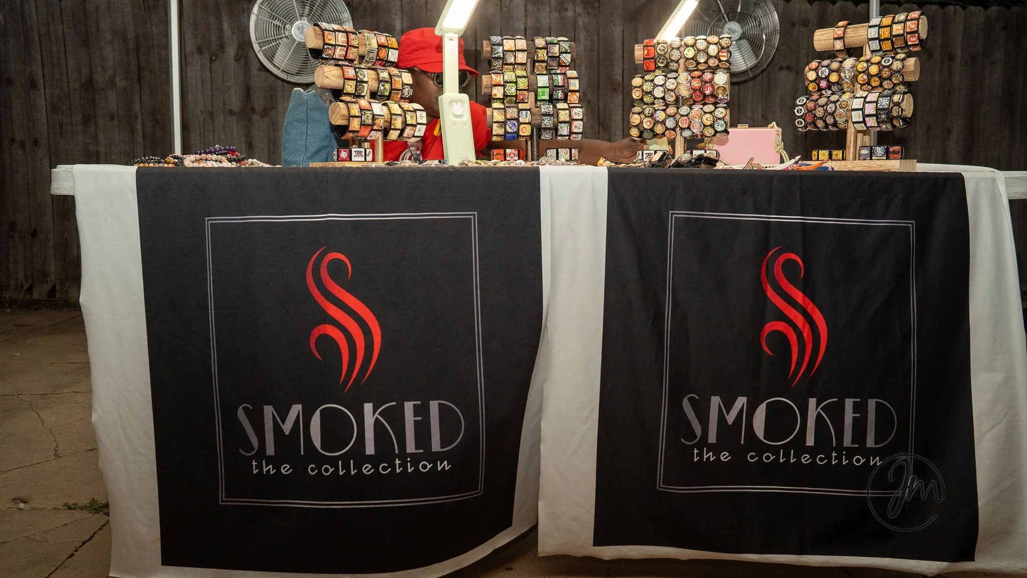A vendor's display at an event featuring rings and jewelry on stands behind a table with black and white tablecloths bearing the logo 'SMOKED the collection' in red and gray. The vendor, partially visible, wears glasses, a red cap, and a red shirt, w