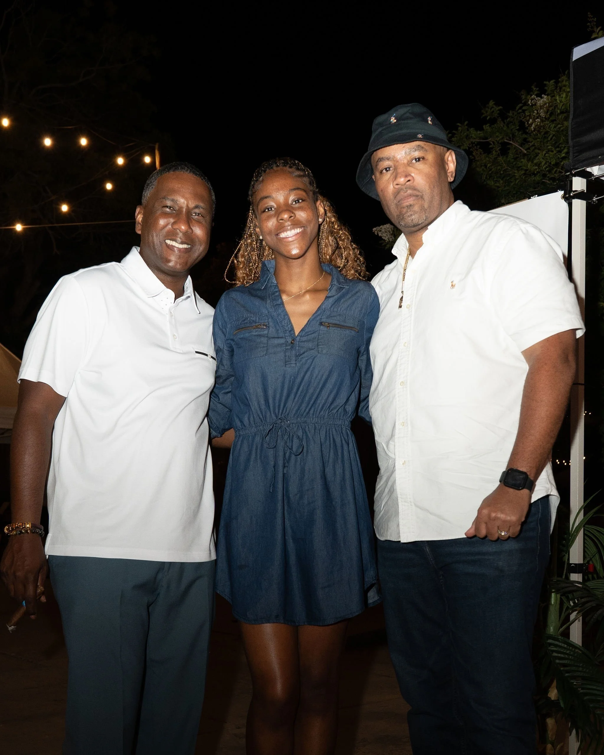 Three people standing together outdoors at night, smiling for the camera, with string lights overhead.