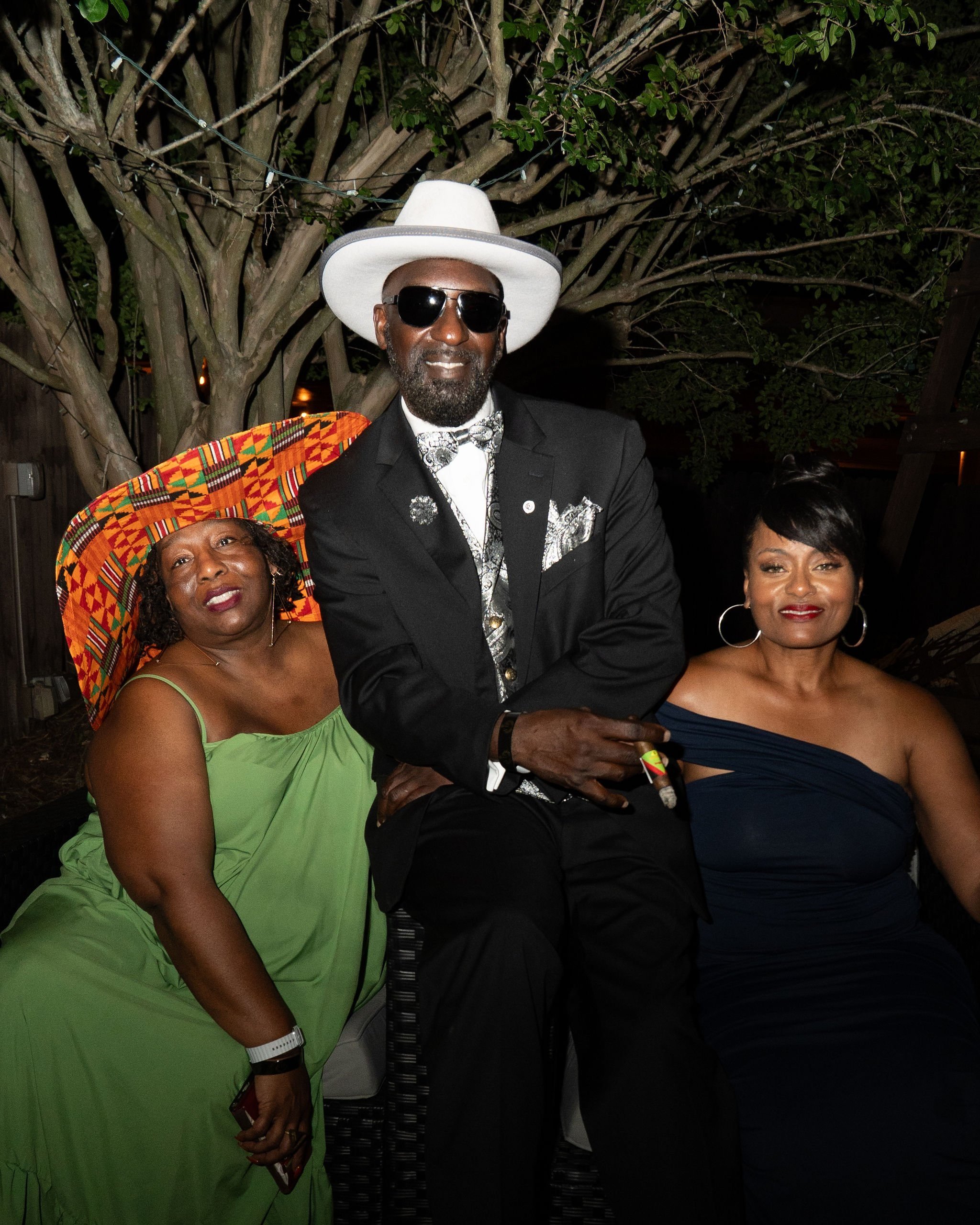 Three people are sitting outdoors at night. The man in the middle is wearing a black suit, a white hat, sunglasses, and a patterned bow tie. The woman on the left is wearing a green dress and a colorful, patterned hat, and the woman on the right is w