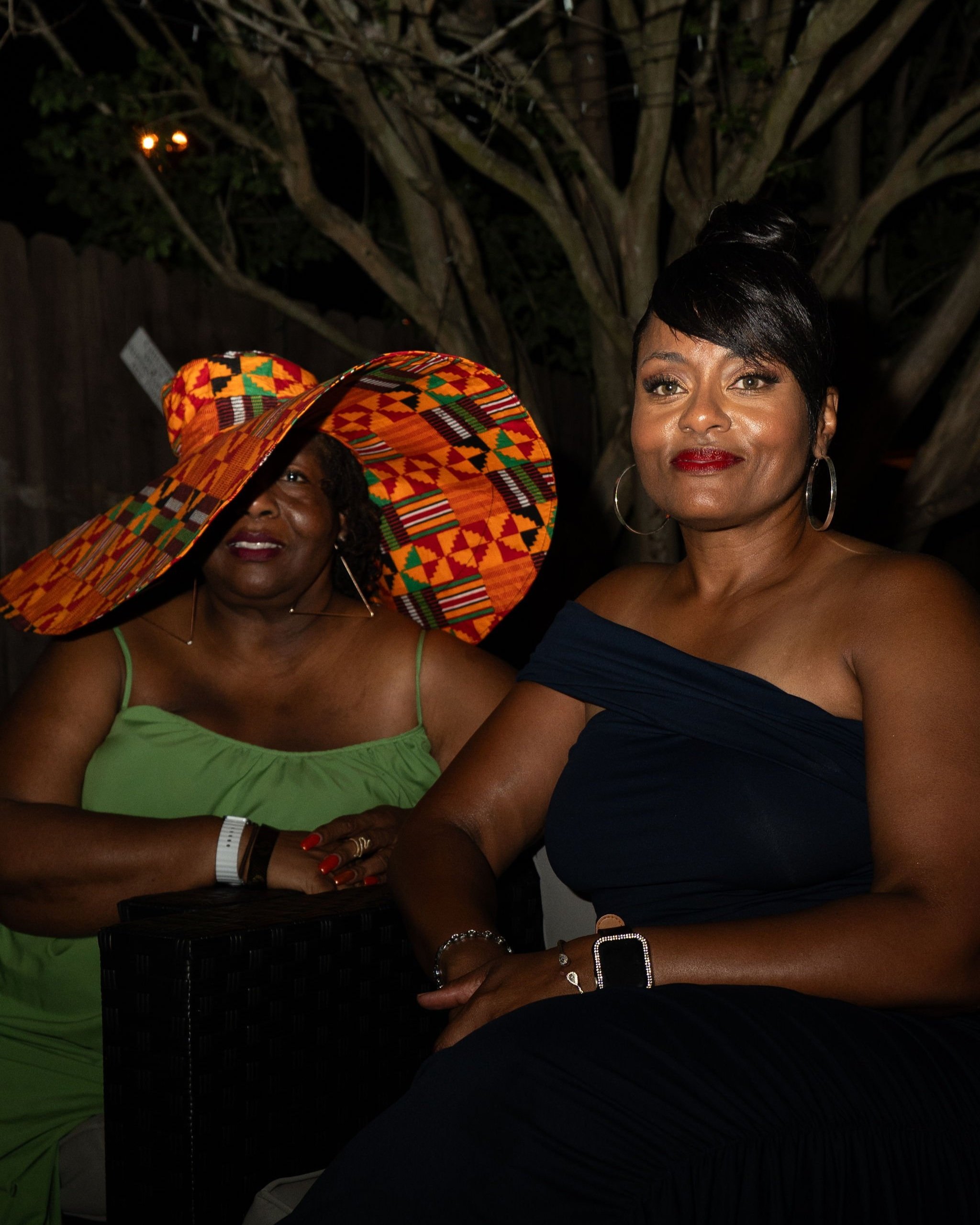 Two women sitting outdoors at night, one wearing a large colorful hat with a Kente pattern, the other in a dark off-shoulder dress, with trees in the background.