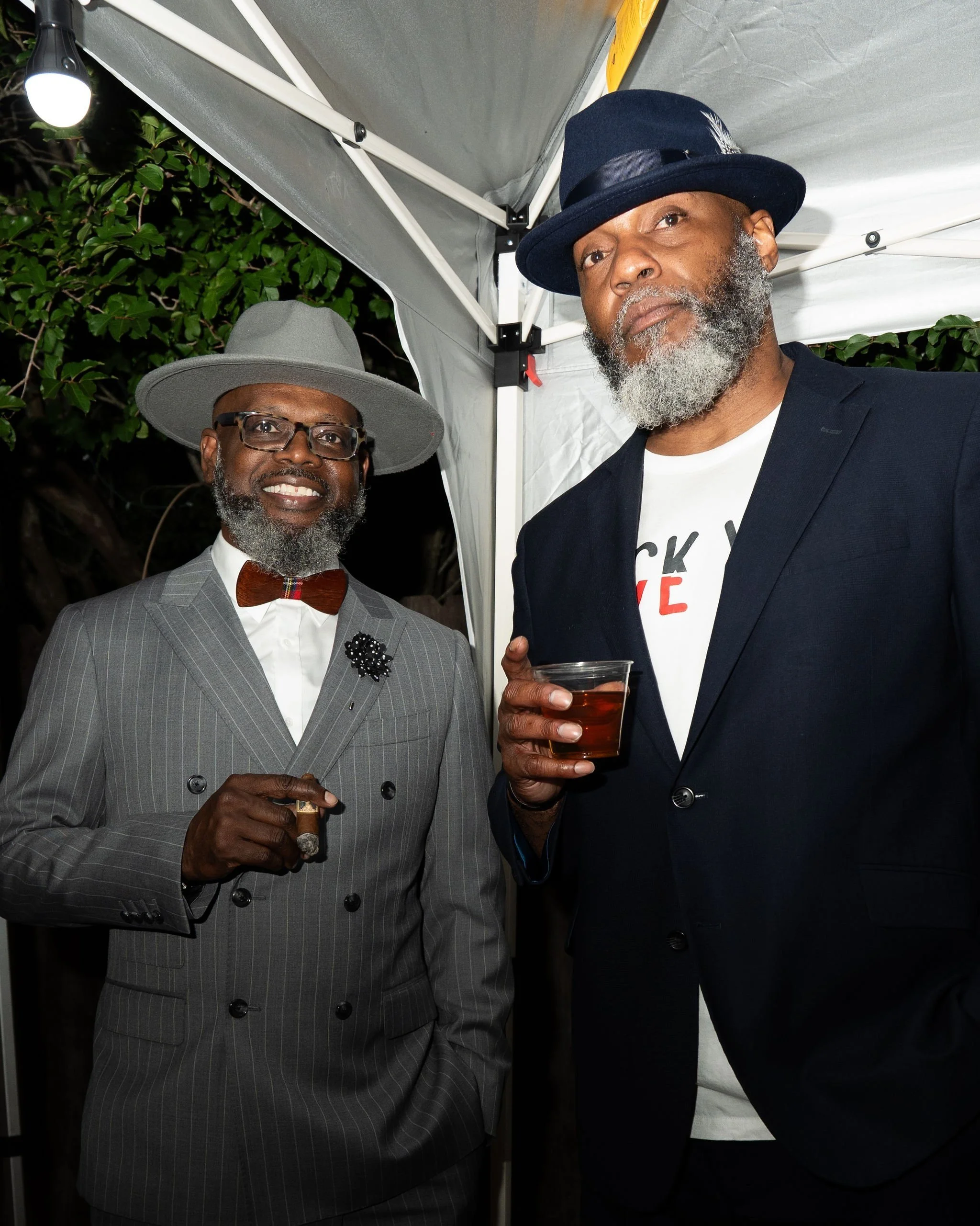 Two stylish men at an outdoor event, one wearing a gray pinstripe suit, gray hat, and glasses, holding a cigar, and the other in a dark blazer, white T-shirt, and a blue hat, holding a drink, standing under a canopy with greenery in the background.