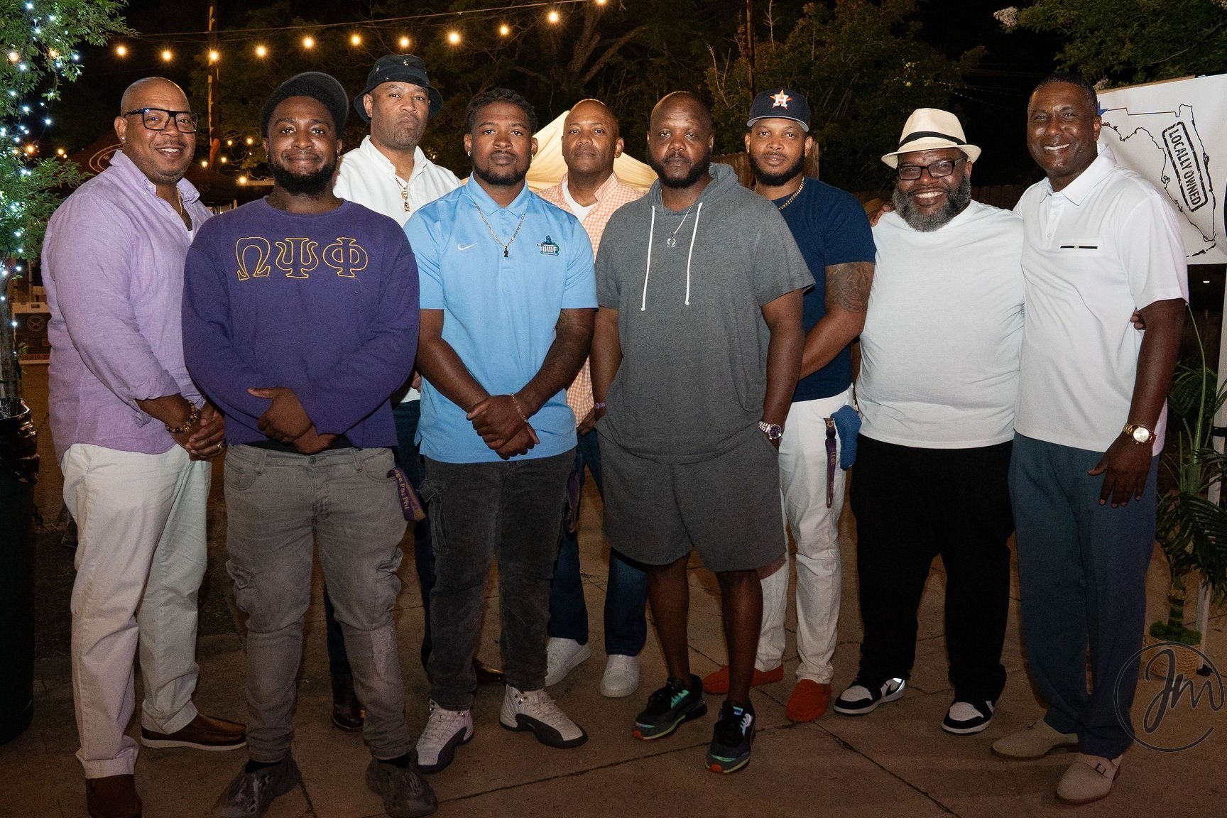 Group of ten men standing outdoors at night, dressed casually, some wearing glasses, hats, or jewelry, with string lights and greenery in the background.