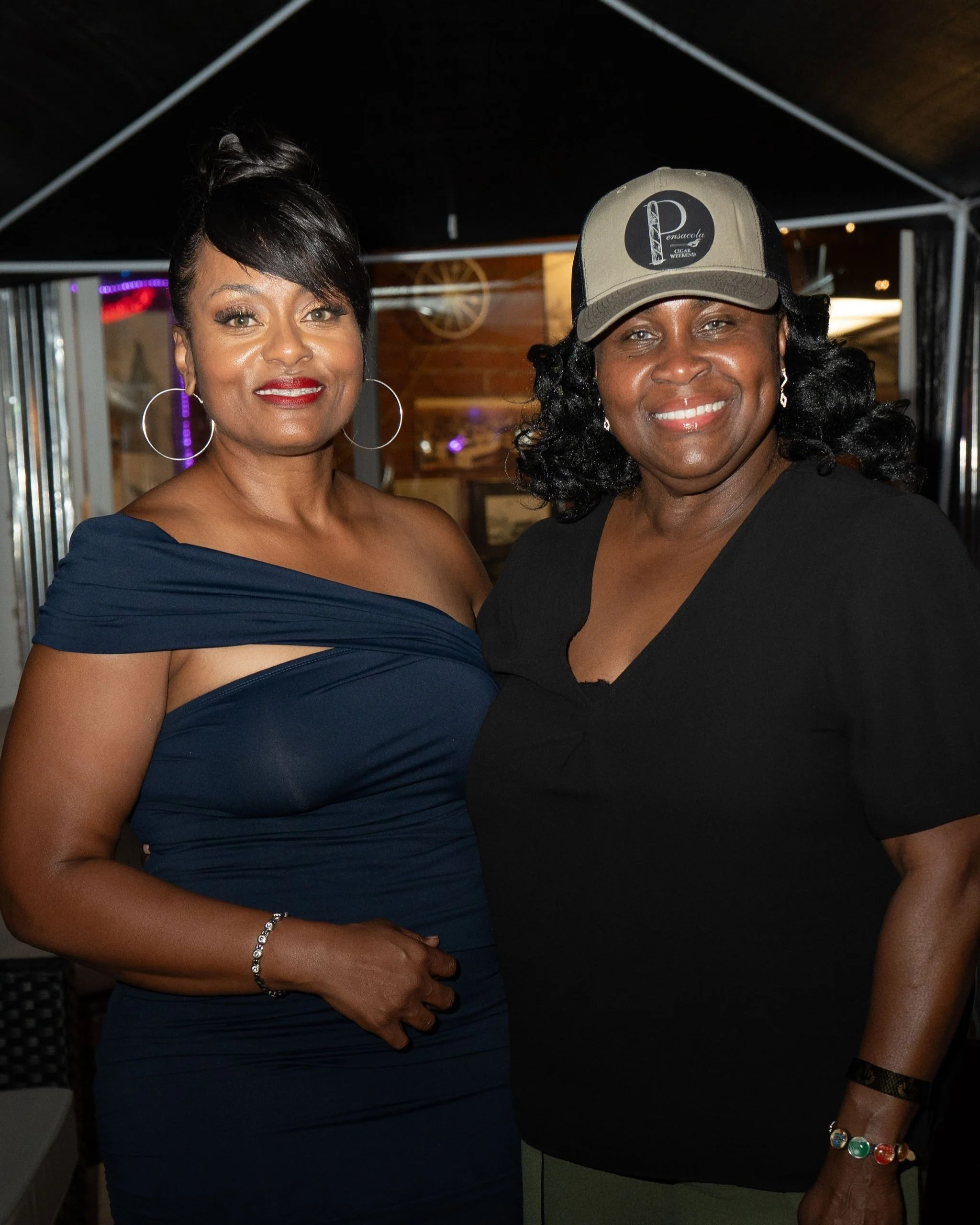 Two women standing side by side, smiling, at an indoor event. The woman on the left is wearing a dark blue off-the-shoulder dress with hoop earrings and a bracelet. The woman on the right is wearing a black shirt, a baseball cap, and a colorful brace