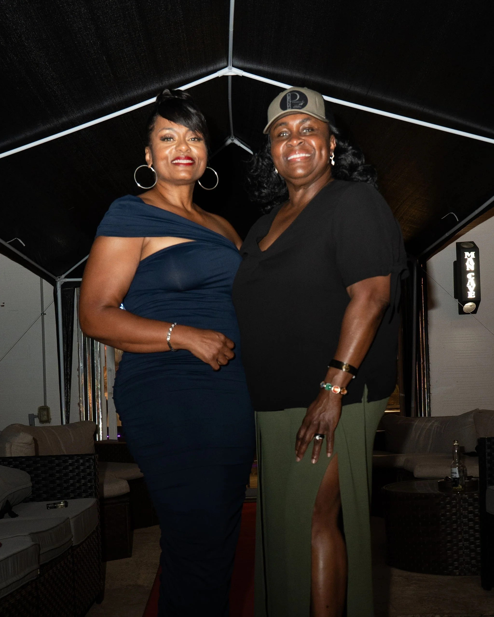 Two women standing close together indoors, smiling at the camera. One is wearing a dark blue dress with an off-shoulder design, and the other is dressed in a black top and olive green skirt with a high slit, accessorized with jewelry and a cap.
