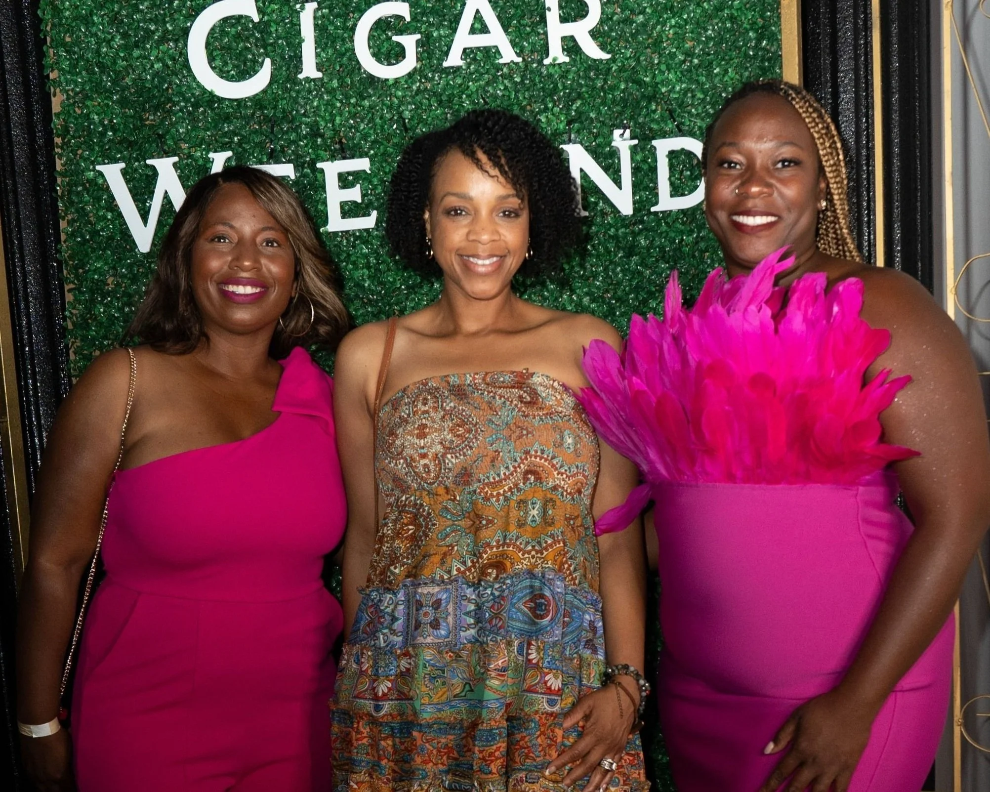 Three women smiling and posing for a photo at an event, with a green leafy backdrop and white letters that spell out 'CIGAR' and other words partially visible.