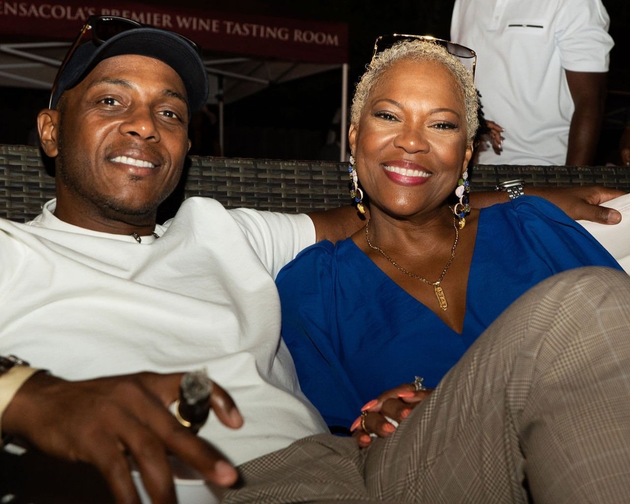 A smiling man and woman sitting close together on a wicker outdoor couch at a wine tasting event, with a dark background and a tent behind them.