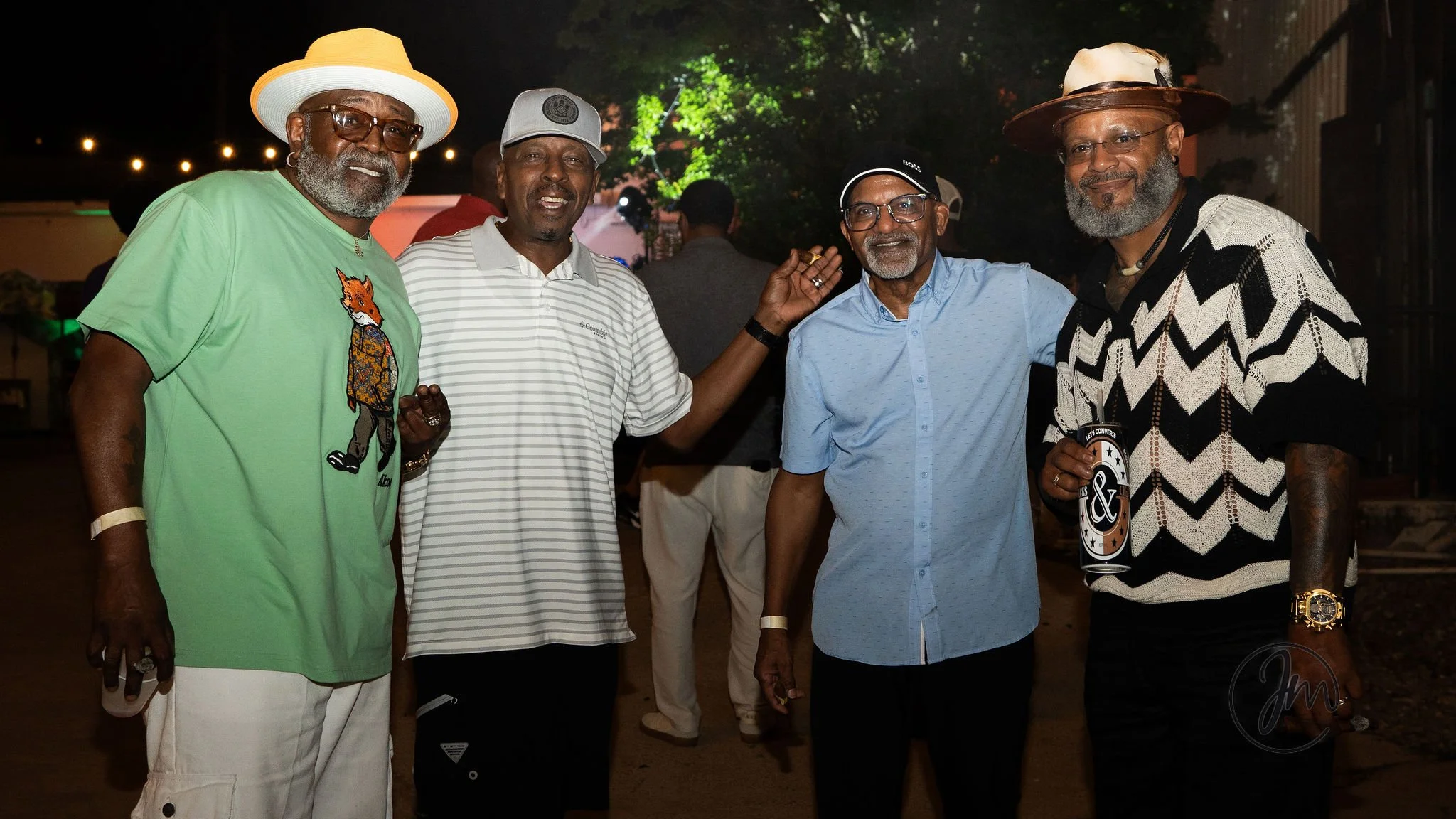 Four older men with gray beards and glasses standing outdoors at night smiling, dressed in casual summer clothing, with string lights and trees in the background.