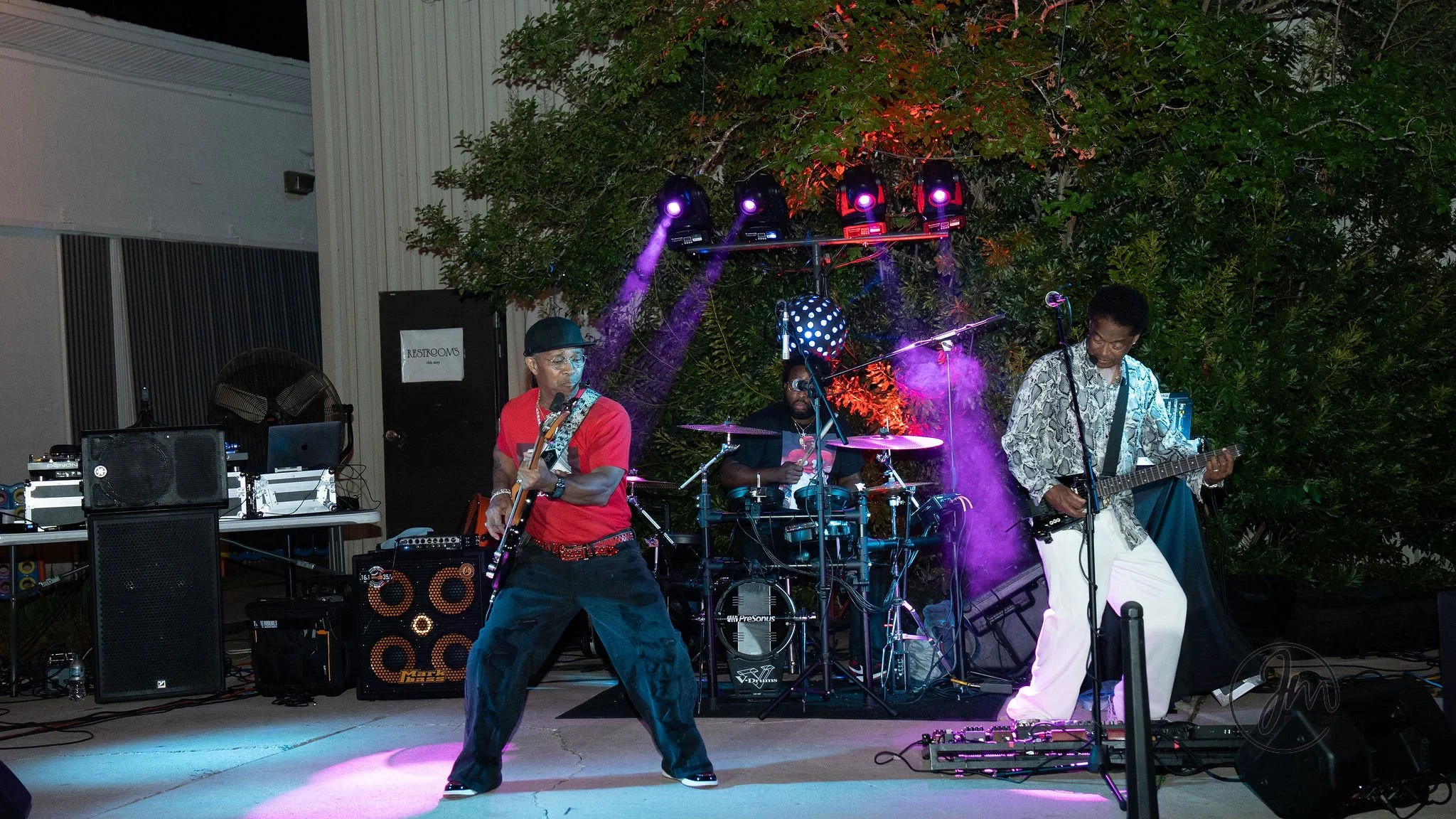 A three-piece band performs on stage outdoors at night with colorful stage lighting, featuring two guitarists and a drummer surrounded by musical equipment.