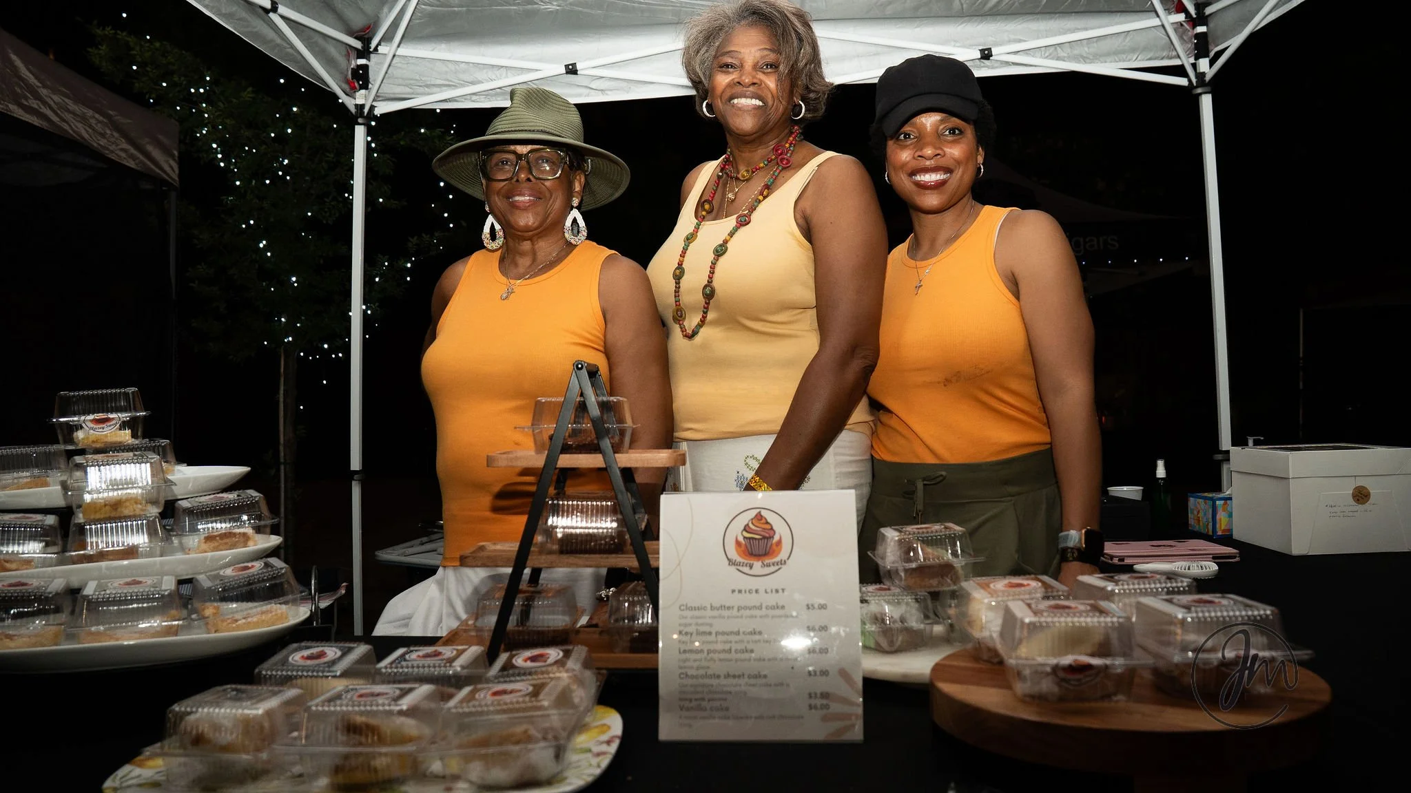 Three women standing behind a table displaying baked goods at a night market booth, smiling for the camera. The table has various boxes of cupcakes and a price list, with a dark outdoor setting and a tent overhead.