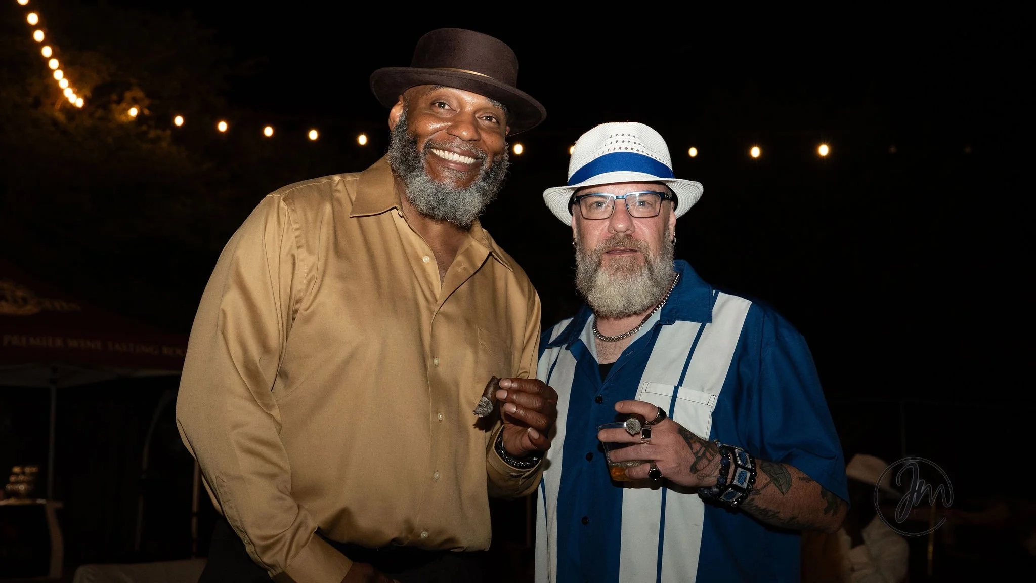 Two middle-aged men with gray beards and glasses at night, one wearing a tan shirt and hat, the other in a blue shirt and white fedora with blue band, standing outdoors under string lights.