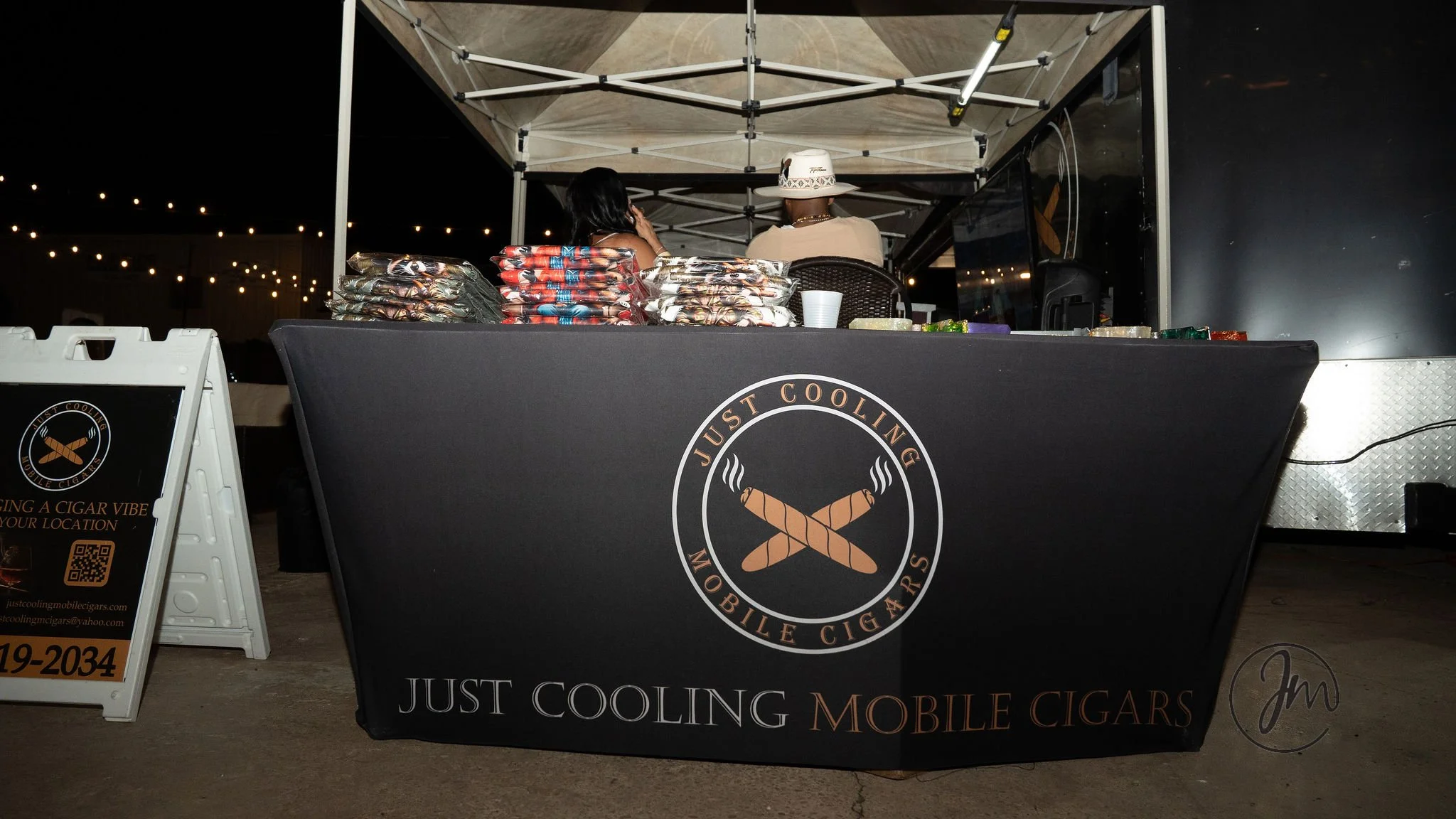 A black vendor booth with a logo for Just Cooling Mobile Cigars, featuring two crossed cigars with steam rising, is set up outdoors at night. There are two people behind the booth, one wearing a white hat, with various packaged cigars and supplies on