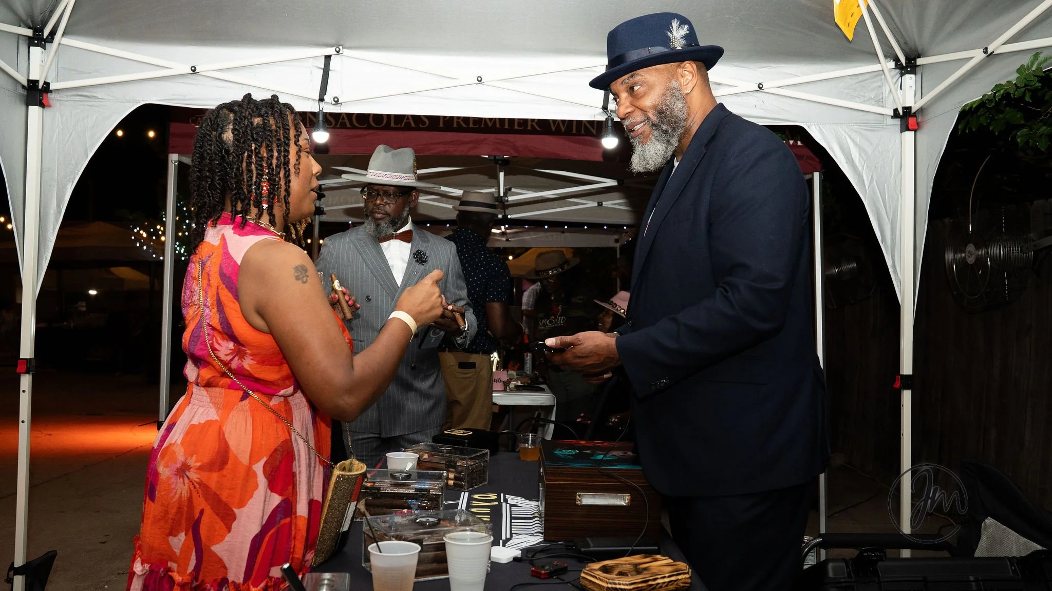 A woman with braids and a colorful dress talking to a man in a black suit and hat at an outdoor event under a white tent, with another man in a gray pinstripe suit and hat in the background.