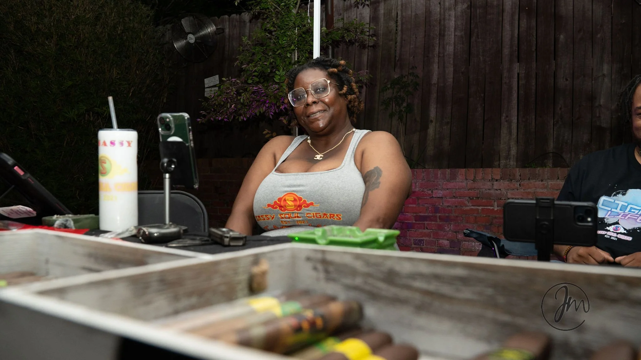 A woman with glasses, wearing a gray tank top with 'Kassy Soul Cigars' logo, sitting at an outdoor table with cigars and a drink, smiling at the camera.