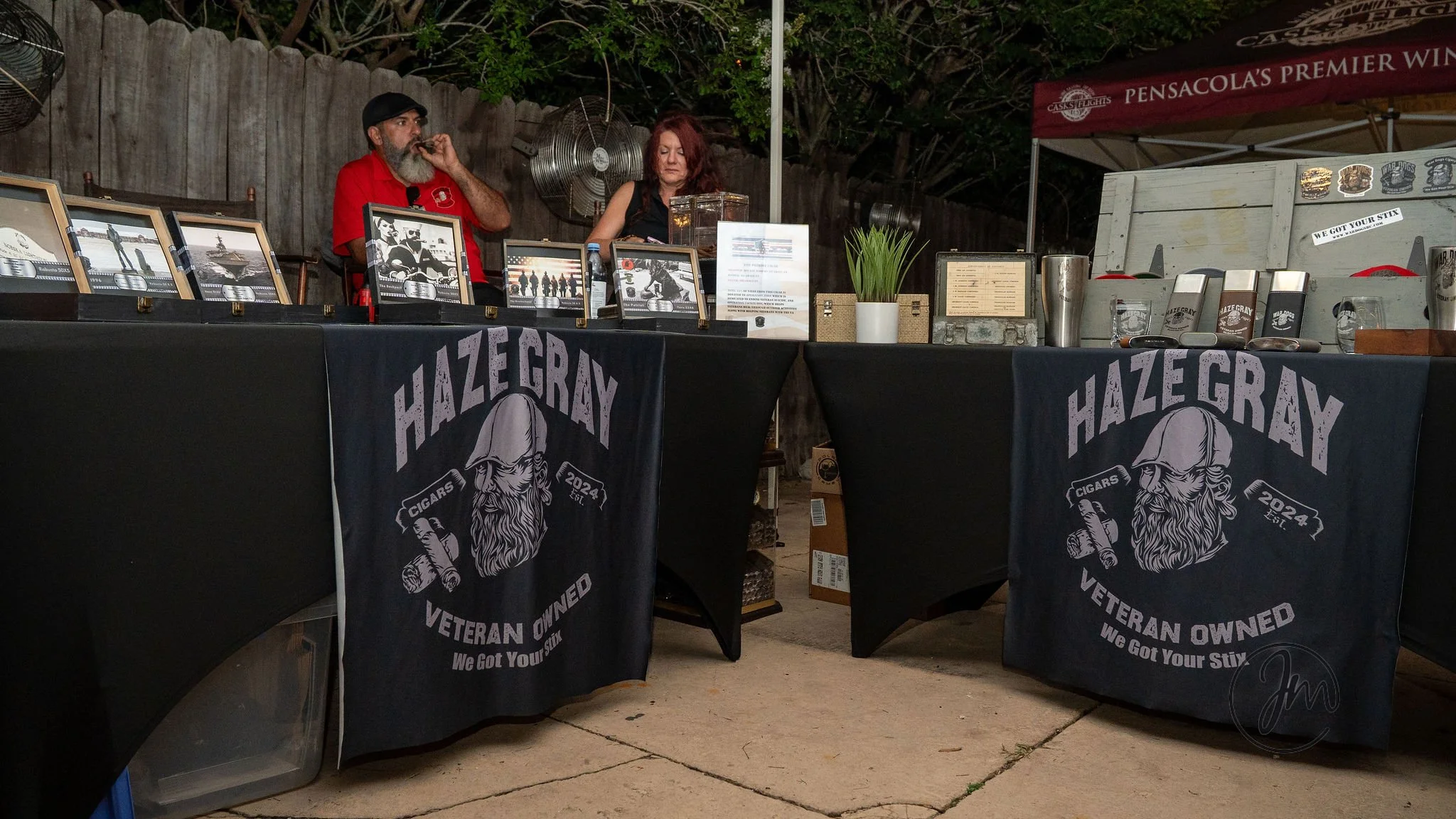 Outdoor display booth with two tables covered by black cloths featuring the Haze Gray logo, a veteran-owned business selling cigars, displaying framed photographs, informational pamphlets, and items like potted plants and metal containers, backed by 