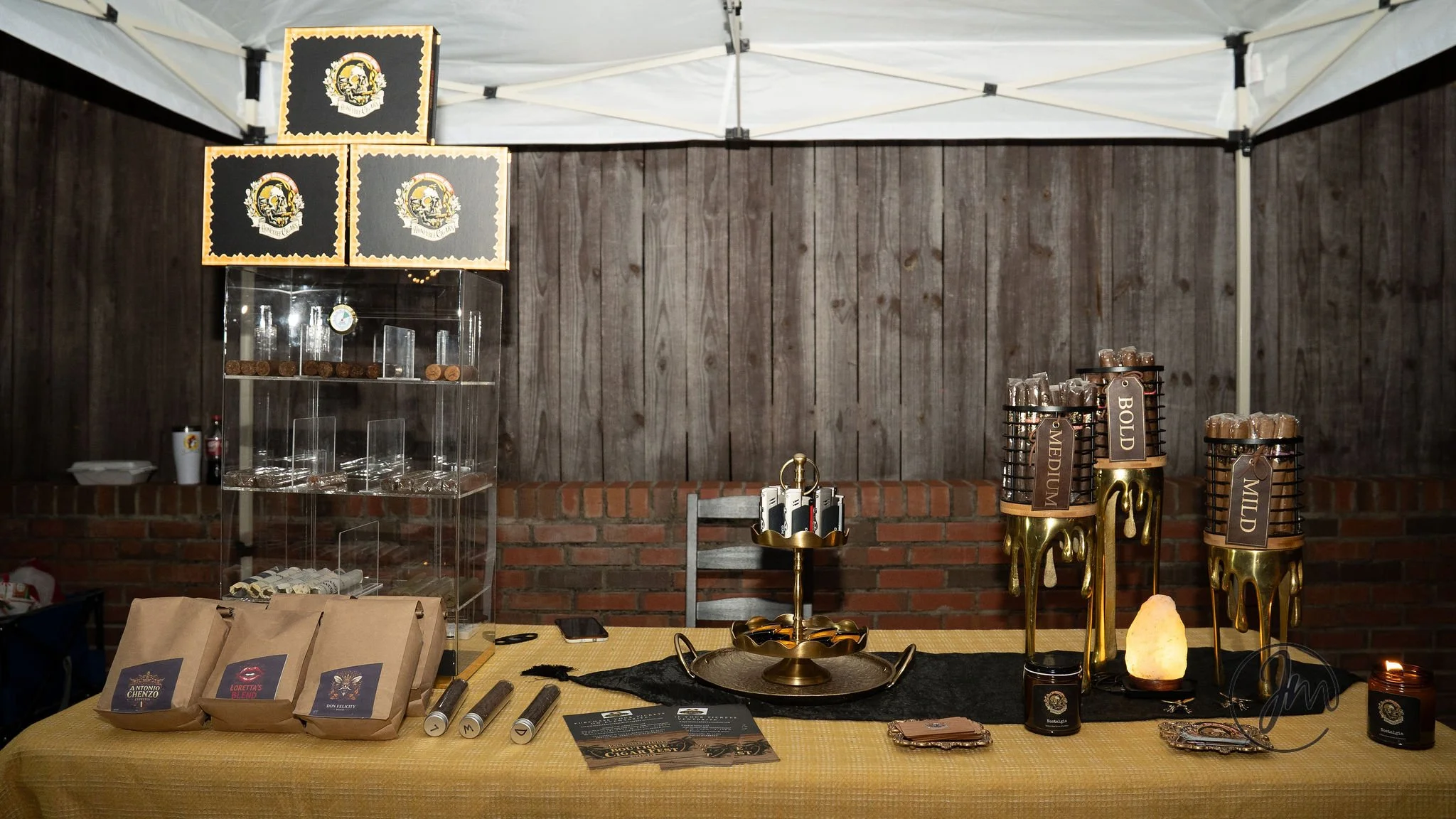 A booth with a yellow tablecloth displaying coffee bags, candles, and drink dispensers. There are decorative signs, glassware, and small items for sale. The background features a wooden wall and a white canopy.