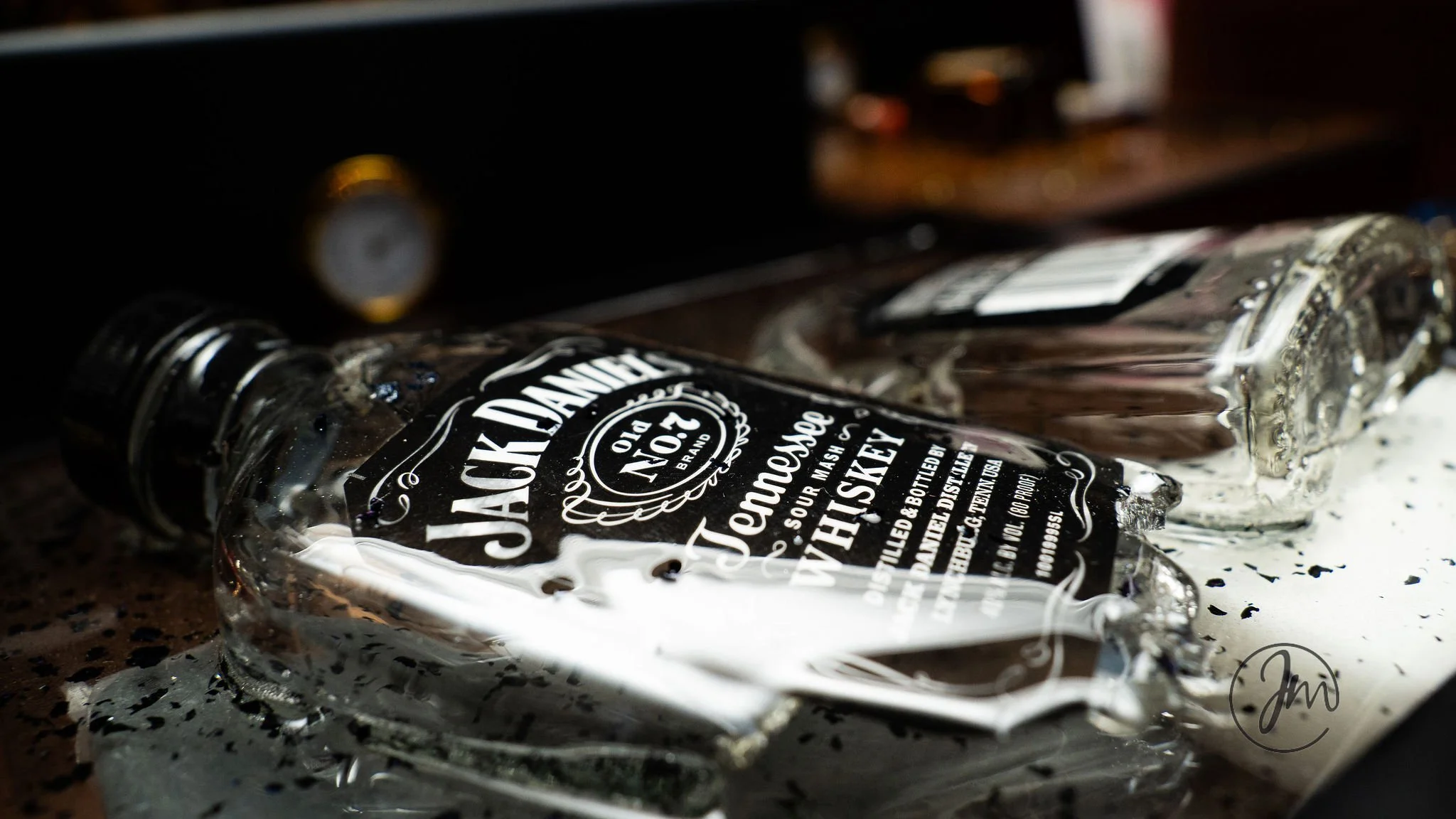 A spilled Jack Daniel's Tennessee Whiskey bottle on a dark surface with a few drops of spilled liquid and water around it.