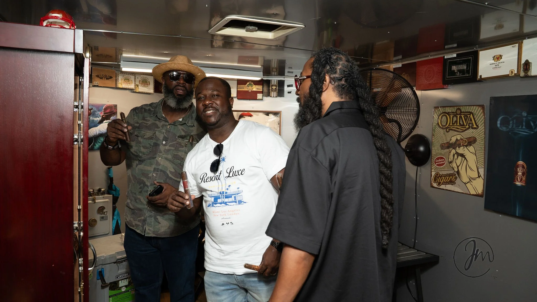 Three men are standing in a room, smoking cigars and having a conversation. The man in the middle is smiling and wearing a white t-shirt with the words 'Resort Luxe' on it. The other two are wearing darker clothing, and one of them is wearing sunglas