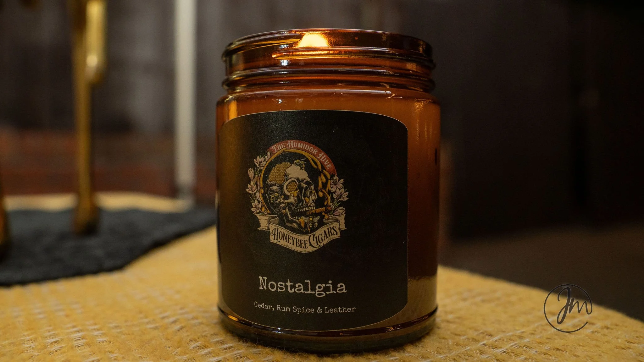 A candle labeled 'Nostalgia' with scents of cedar, rum spice, and leather, from Honeybee Cigars, on a textured surface with a blurred background.