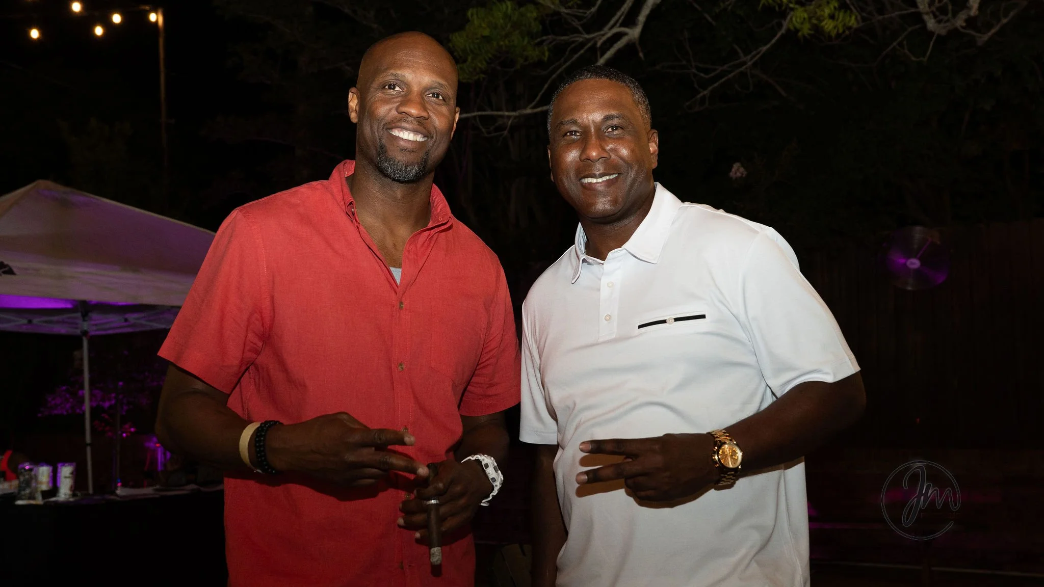 Two men standing outdoors at night, smiling, with one wearing a red shirt and the other wearing a white shirt, both flashing peace signs.
