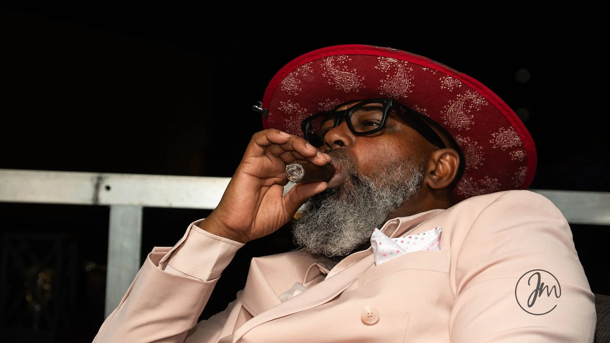 A man with a gray beard and glasses, wearing a pink suit and a red wide-brimmed hat, is smoking a cigar.