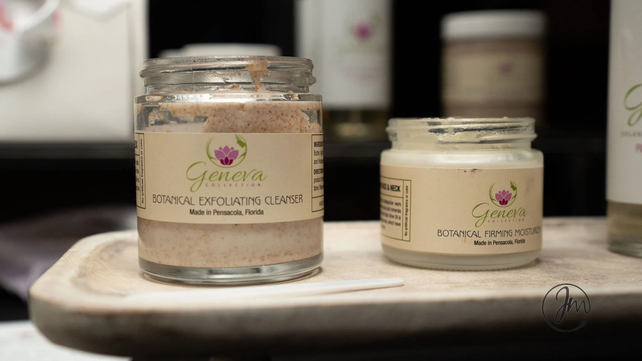 Close-up of two cosmetic jars on a wooden surface, one labeled 'Botanical Exfoliating Cleanser' and the other 'Botanical Firming Moisturizer,' both from Geneva Collection, made in Pensacola, Florida.