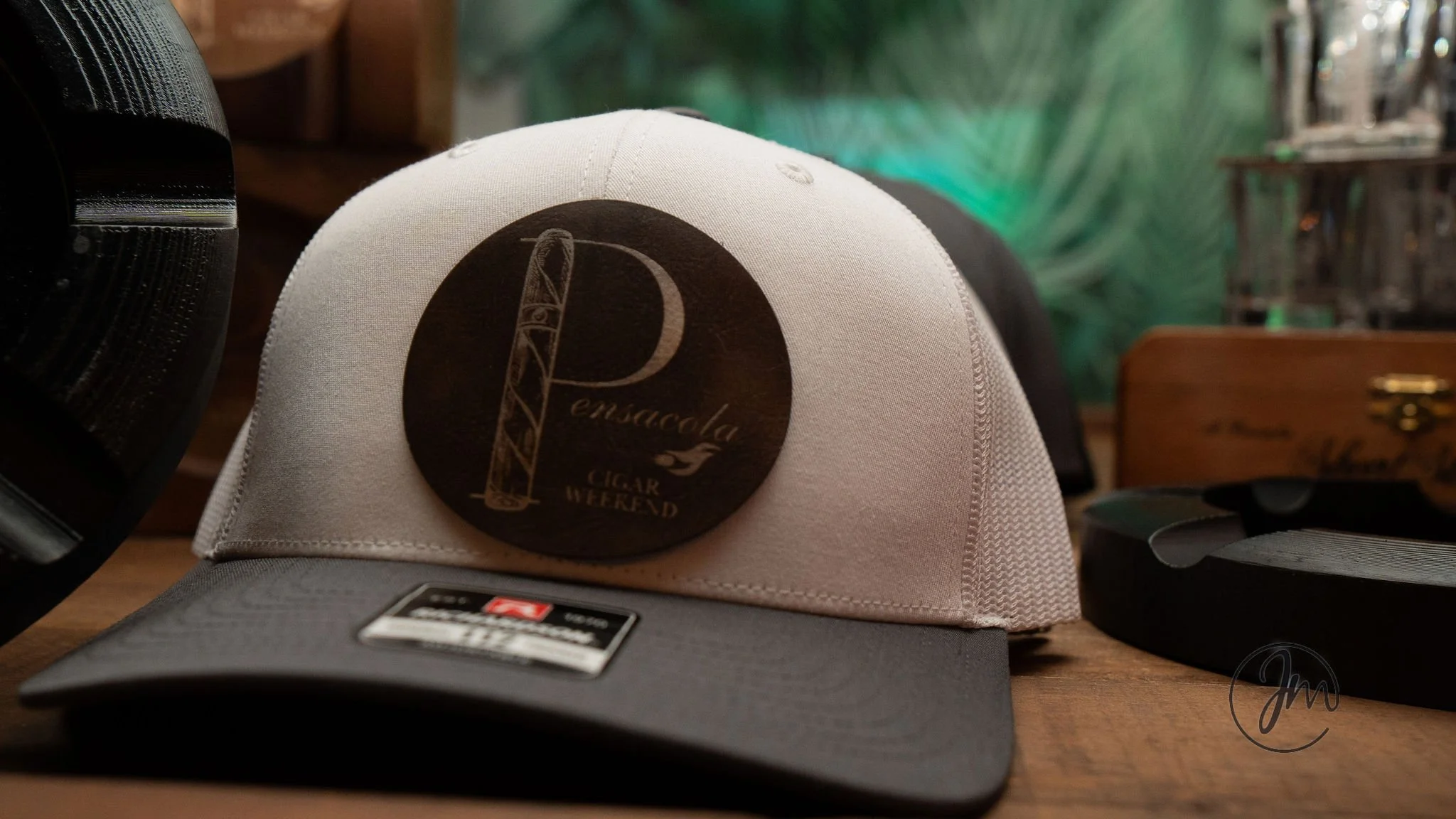 White and black baseball cap with a round black sticker that reads 'Pensacola Cigar Weekend' on the front, placed on a wooden surface alongside other items.