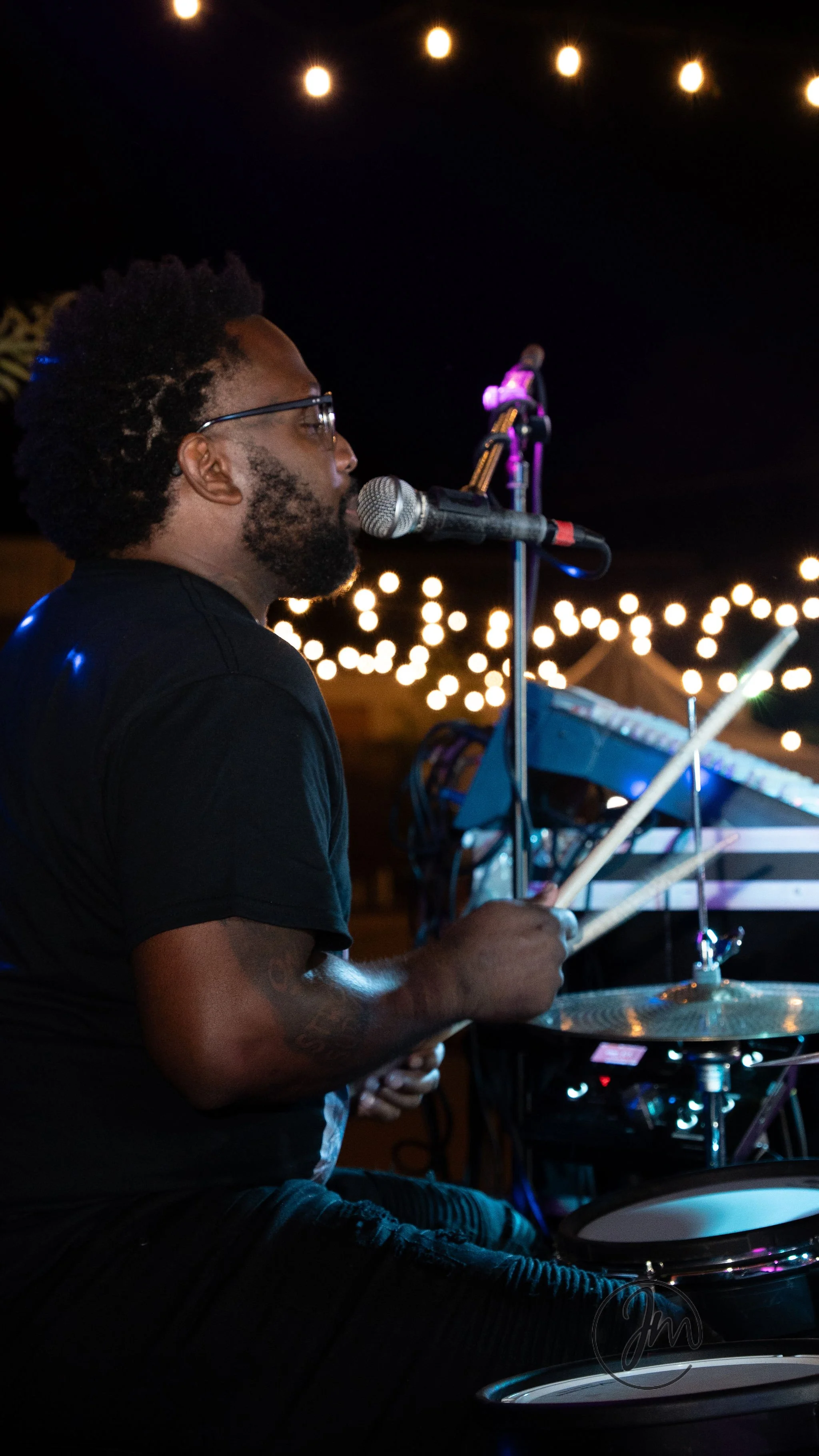 A man with glasses and a beard playing drums at night under string lights, with a microphone nearby.