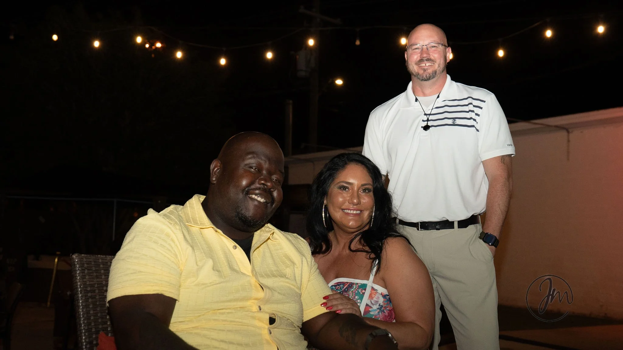 Three adults, two men and one woman, smiling at an outdoor evening gathering with string lights overhead. The man on the left is wearing a yellow shirt, the woman in the middle is wearing a colorful strapless dress, and the man on the right is dresse
