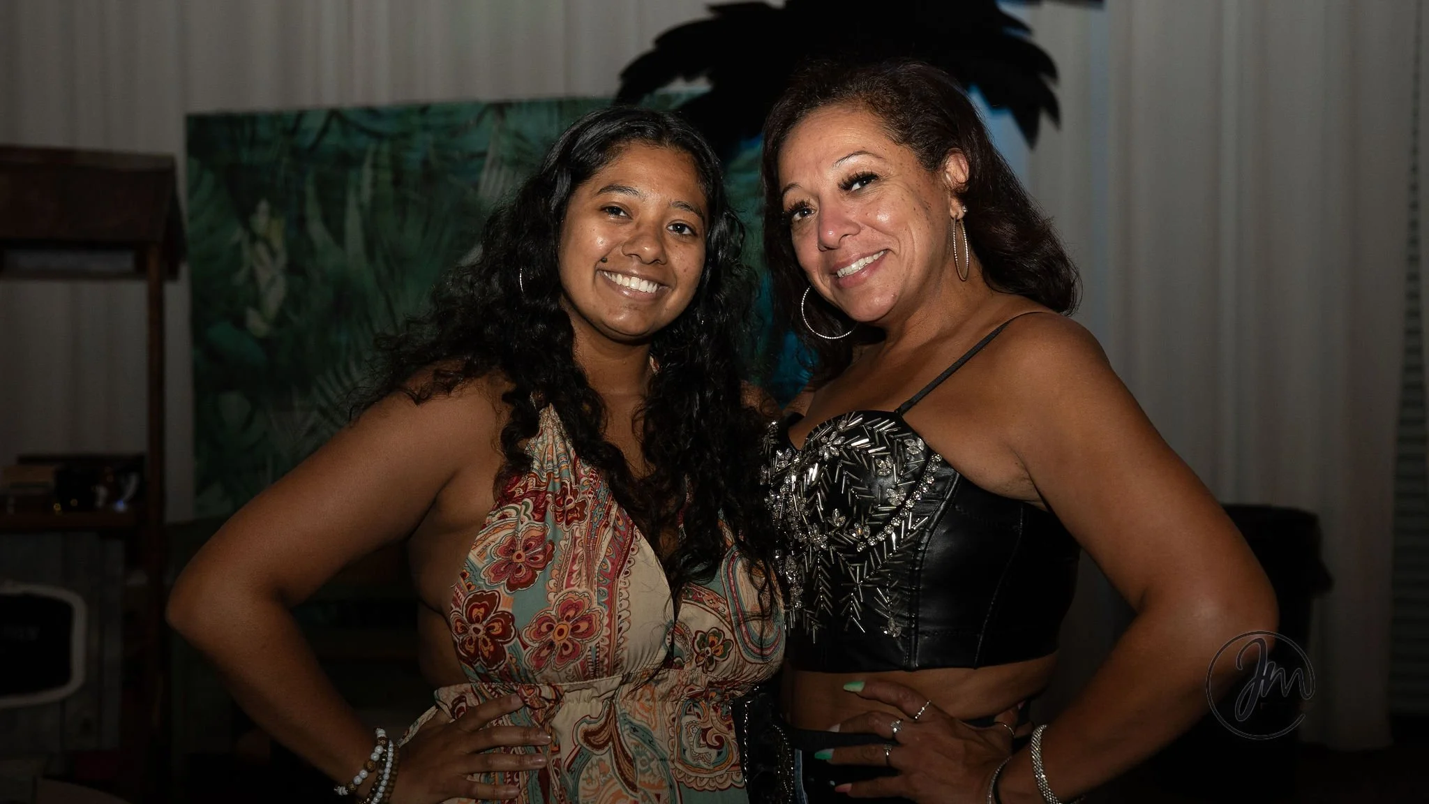 Two women smiling and posing together indoors, one with long curly hair wearing a colorful patterned dress, and the other with short dark hair wearing a black leather top with silver embellishments.
