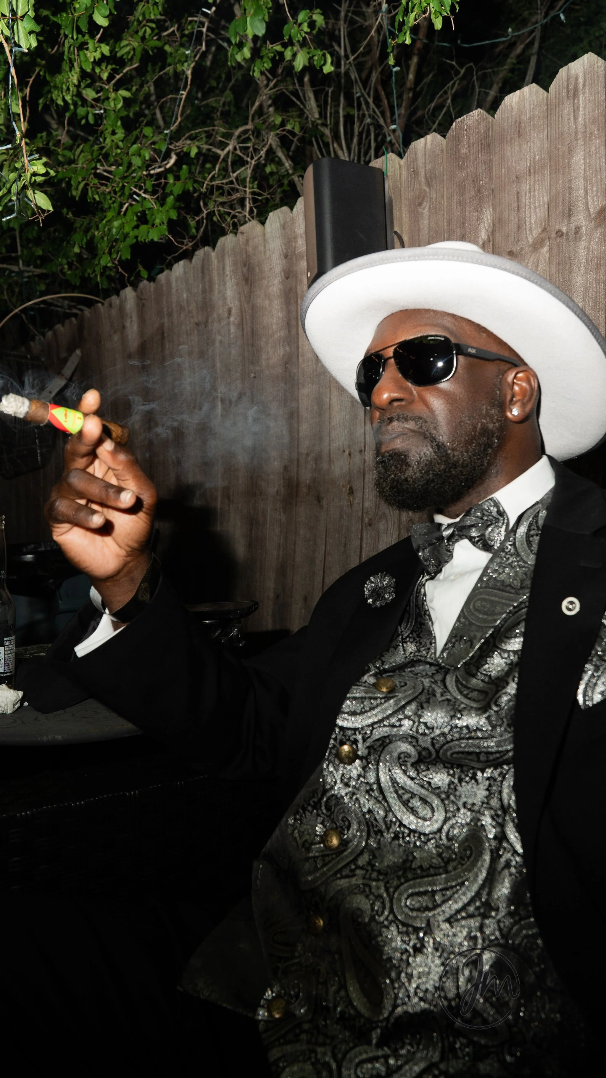 A man dressed in formal attire with a paisley vest, black suit, bow tie, sunglasses, and a large white hat, sitting outdoors, smoking a cigar.