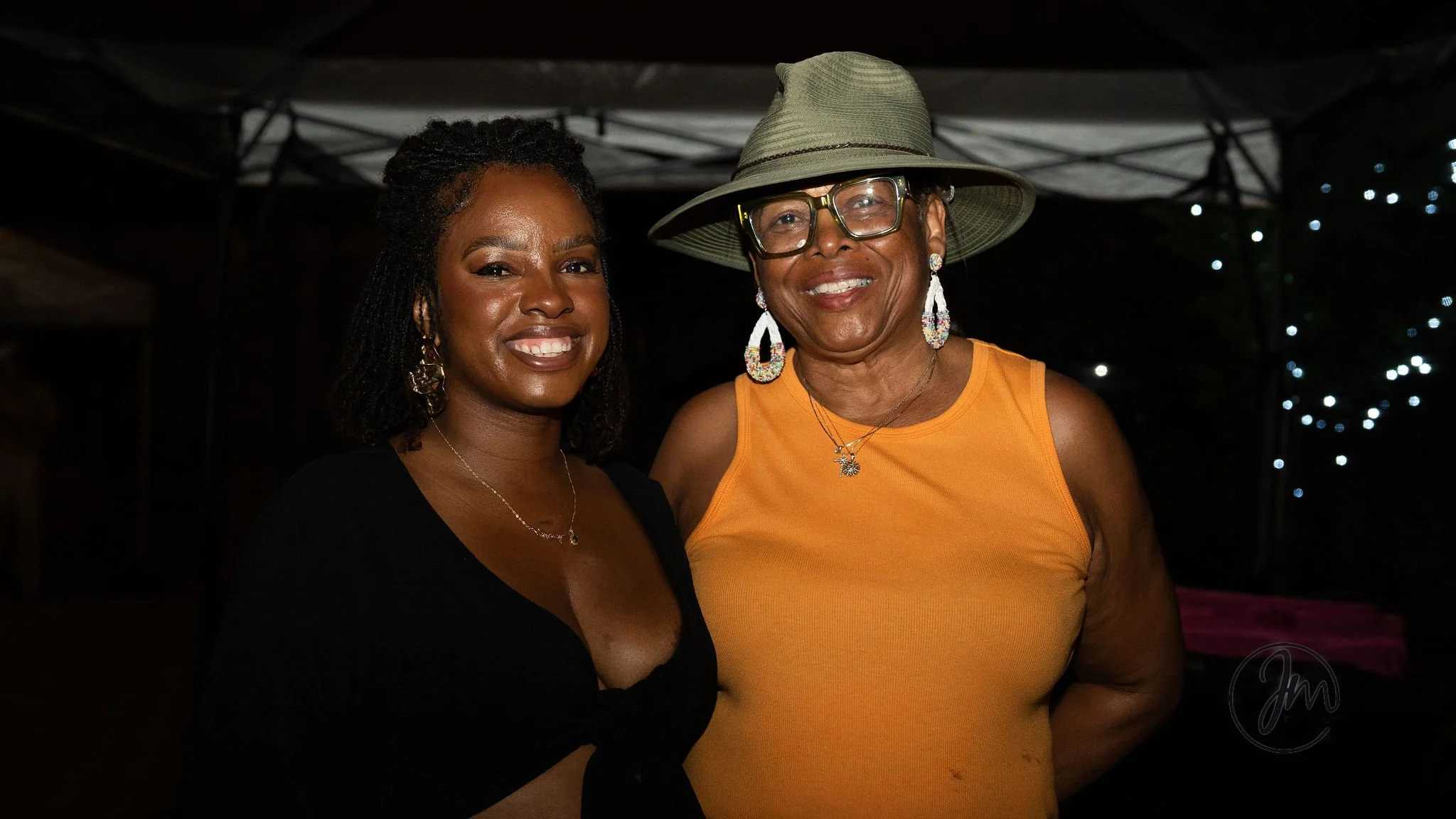 Two women standing close together, smiling, at night with string lights in the background; one woman wears a black top, and the other wears an orange sleeveless shirt, large hat, glasses, and jewelry.