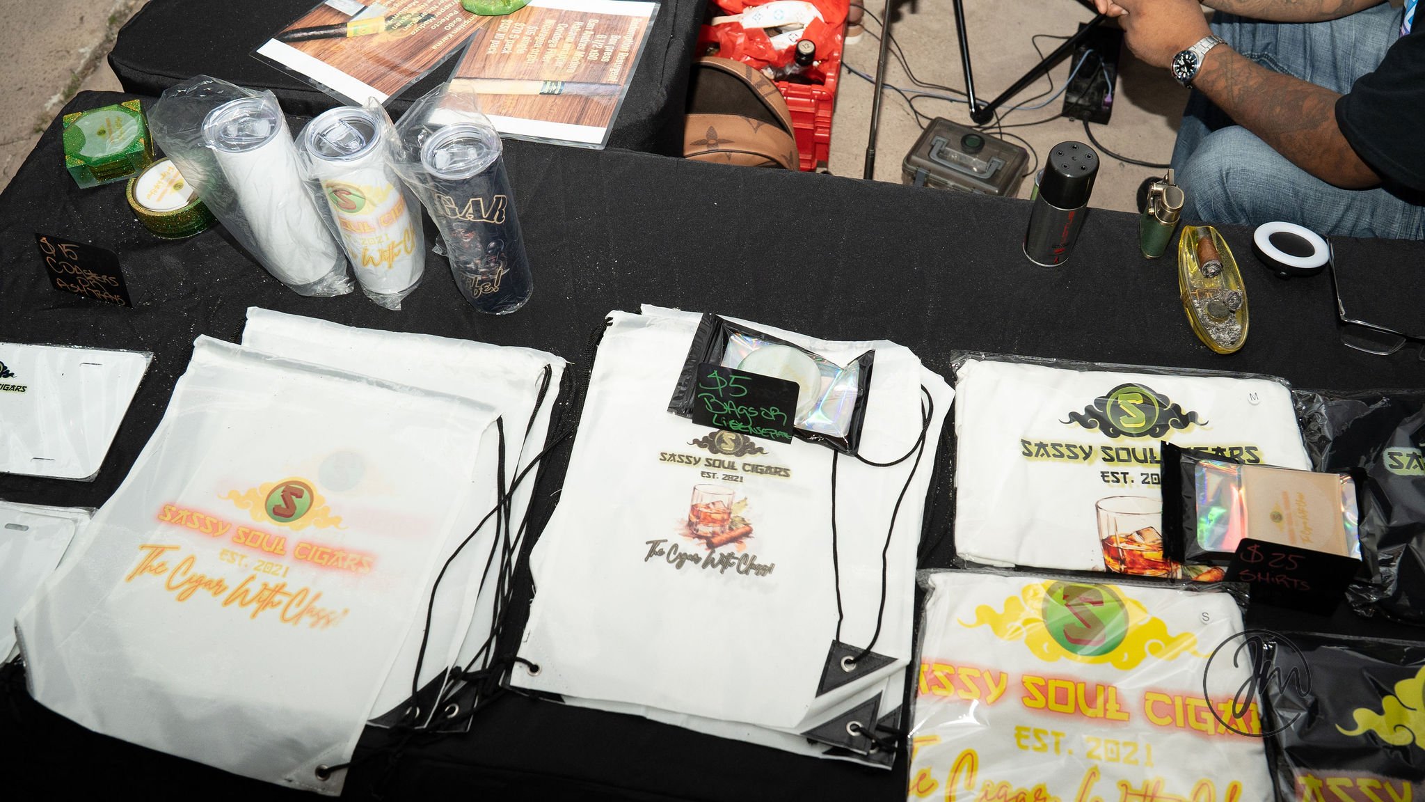 A display table at Sassy Soul Cigars with branded white paper bags, some containing cigars, and various vaping or smoking accessories. The table has a black cloth, and there are drinks, a lighter, and other items visible.