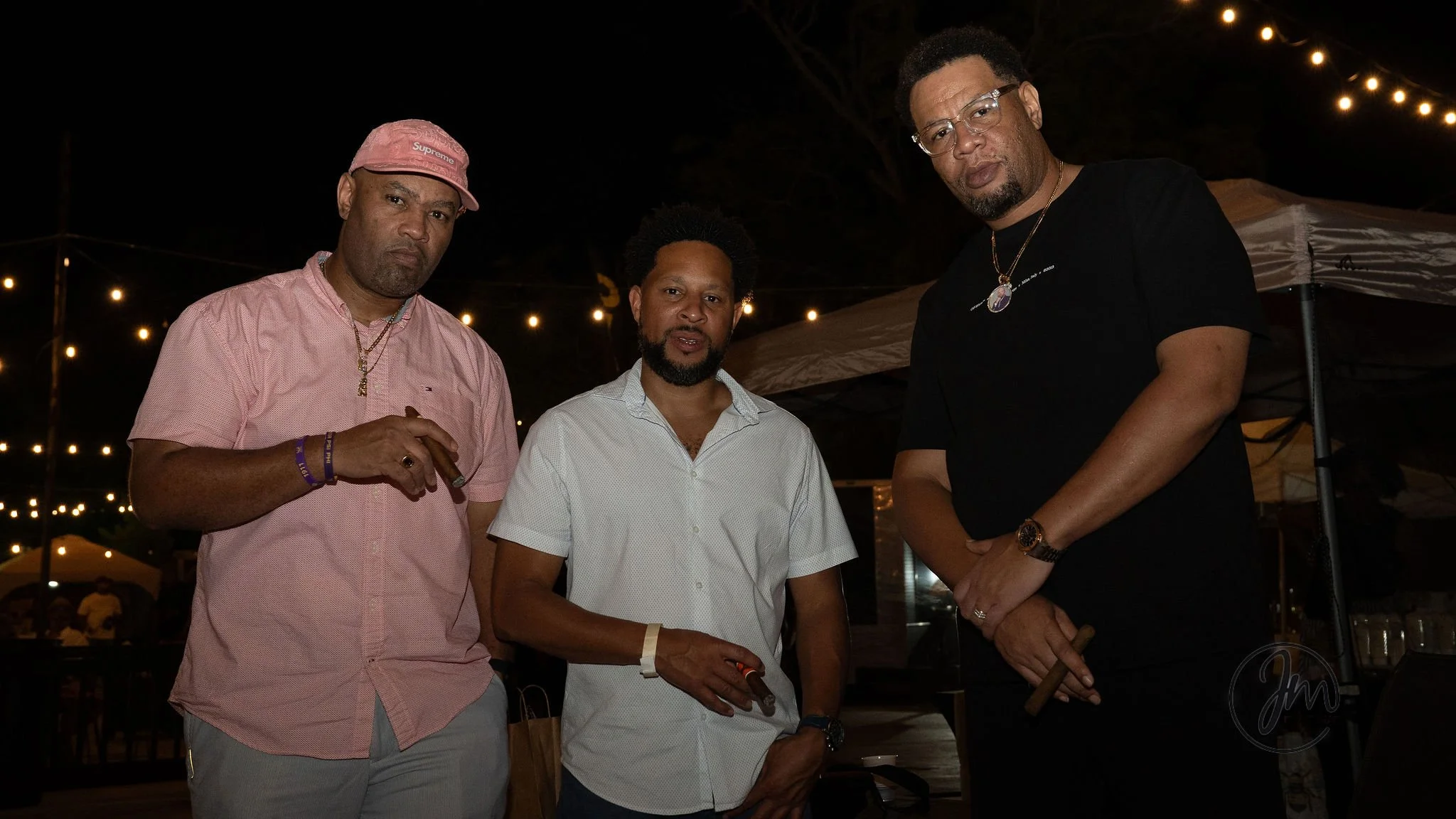Three men standing together at an outdoor event at night, illuminated by string lights, with tents in the background.