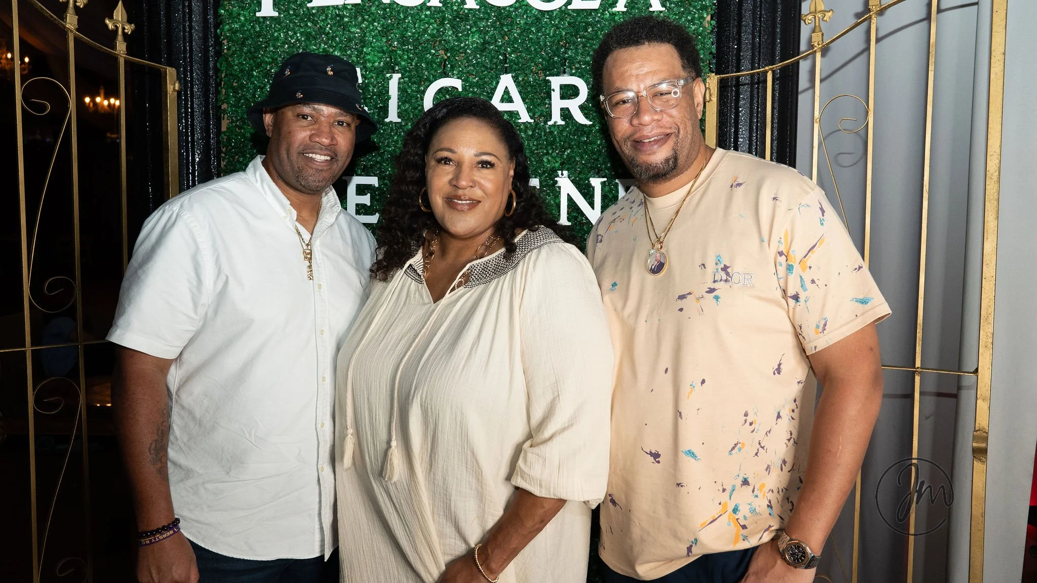 Three people standing together smiling at an event with a green foliage backdrop and gold gate, one man on the left wearing a white shirt and black bucket hat, woman in the middle in a cream dress, man on the right in a beige tie-dye shirt with jewel