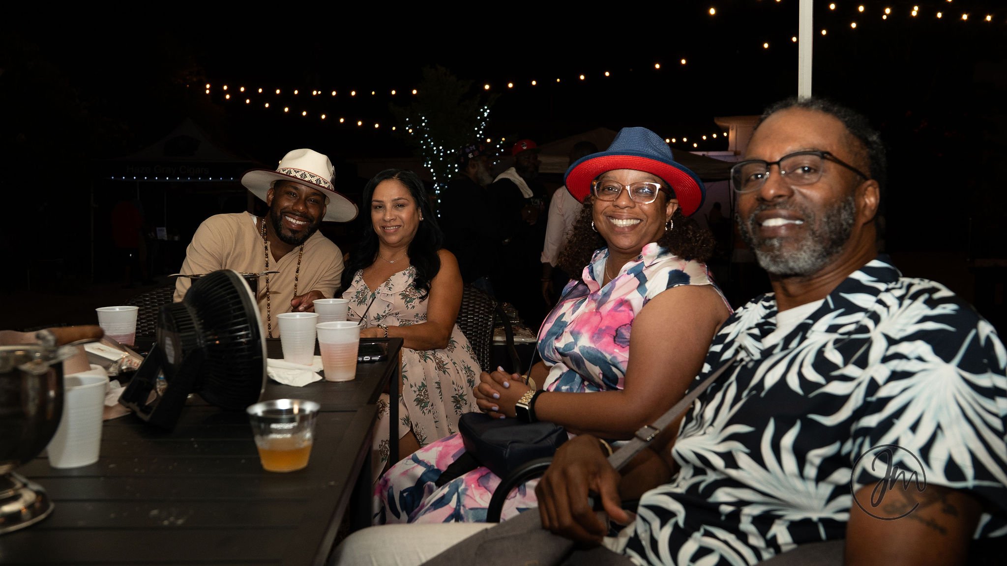 Four adults sitting at a table outdoors at night, smiling for the camera, under string lights. One man wears a white hat, and a woman wears a colorful hat and floral dress. The table has cups and drinks on it.