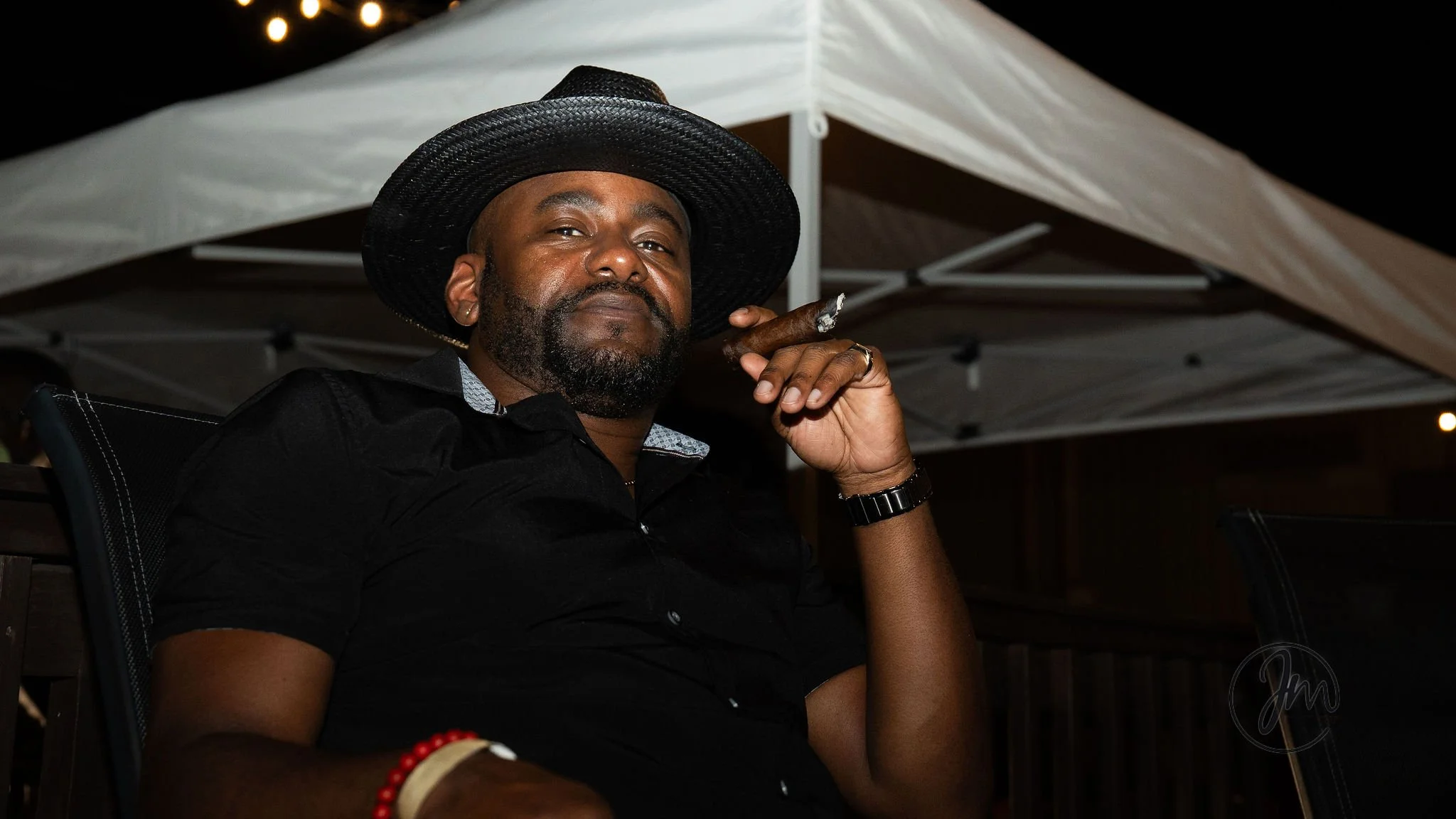 A man with a beard wearing a black shirt, a large black hat, and a watch, sitting outdoors at night under a canopy while holding a cigar and looking at the camera.