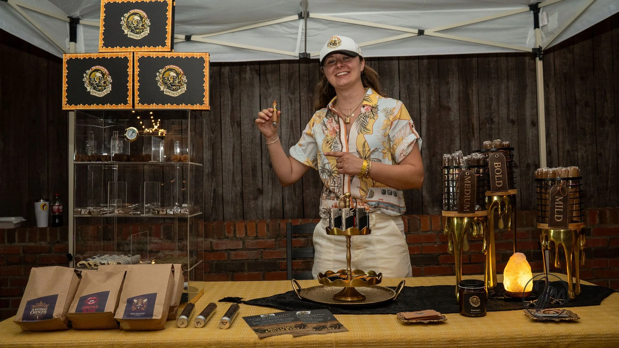 Woman smiling at a cigar booth, holding a cigar and pointing at it. The booth has cigar samples, bags, and jars labeled 'JMEDIUM,' 'BOLD,' and 'MILD.' There are also decorative items including a salt lamp and framed signs with a skull logo.
