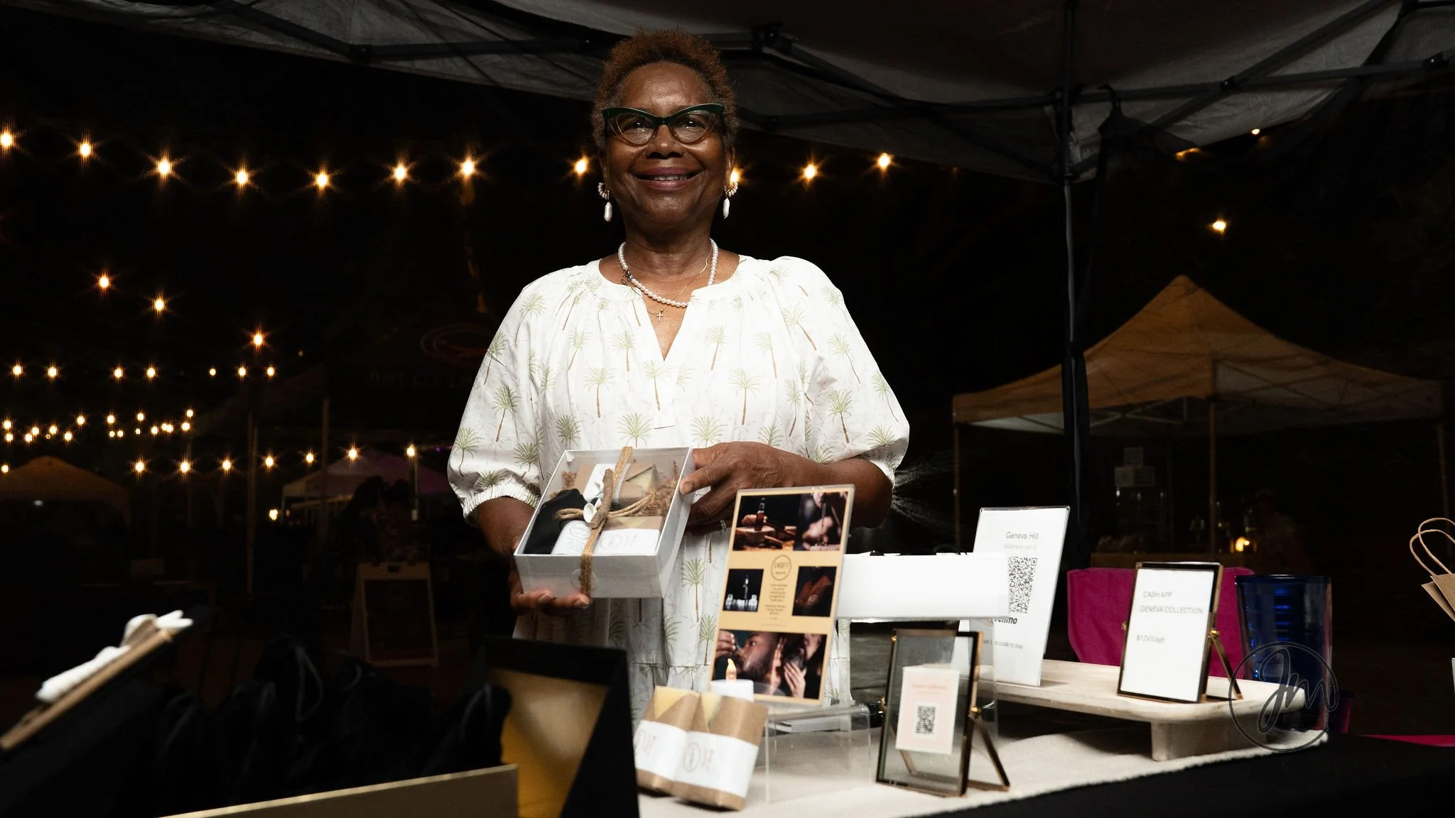 An older woman with glasses and jewelry stands behind a table at an outdoor market, holding a box of items on display with string decorations, illuminated by string lights at night.