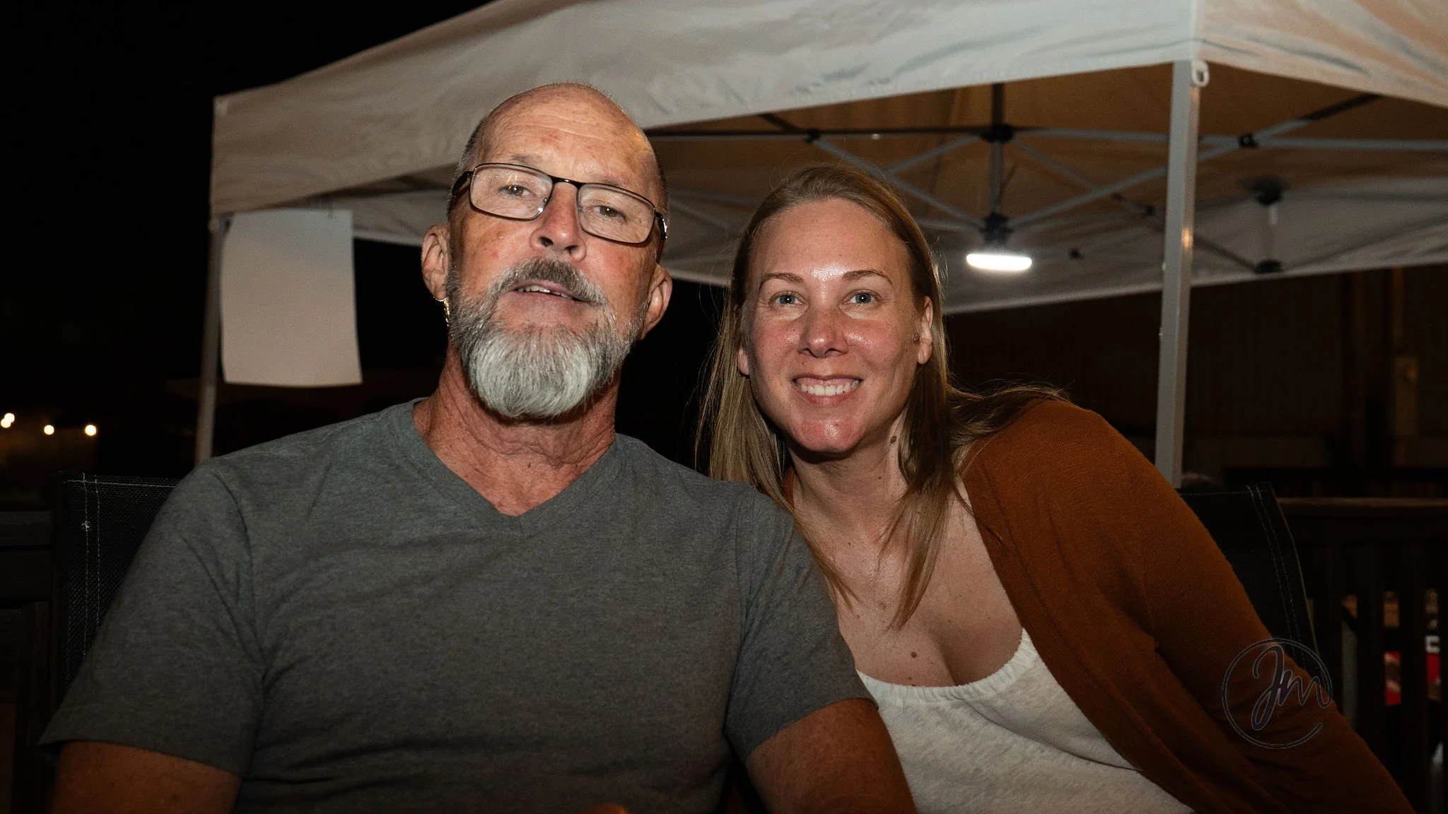 A smiling man with a beard and glasses sitting next to a smiling woman with red hair at night at an outdoor setting with a canopy tent in the background.