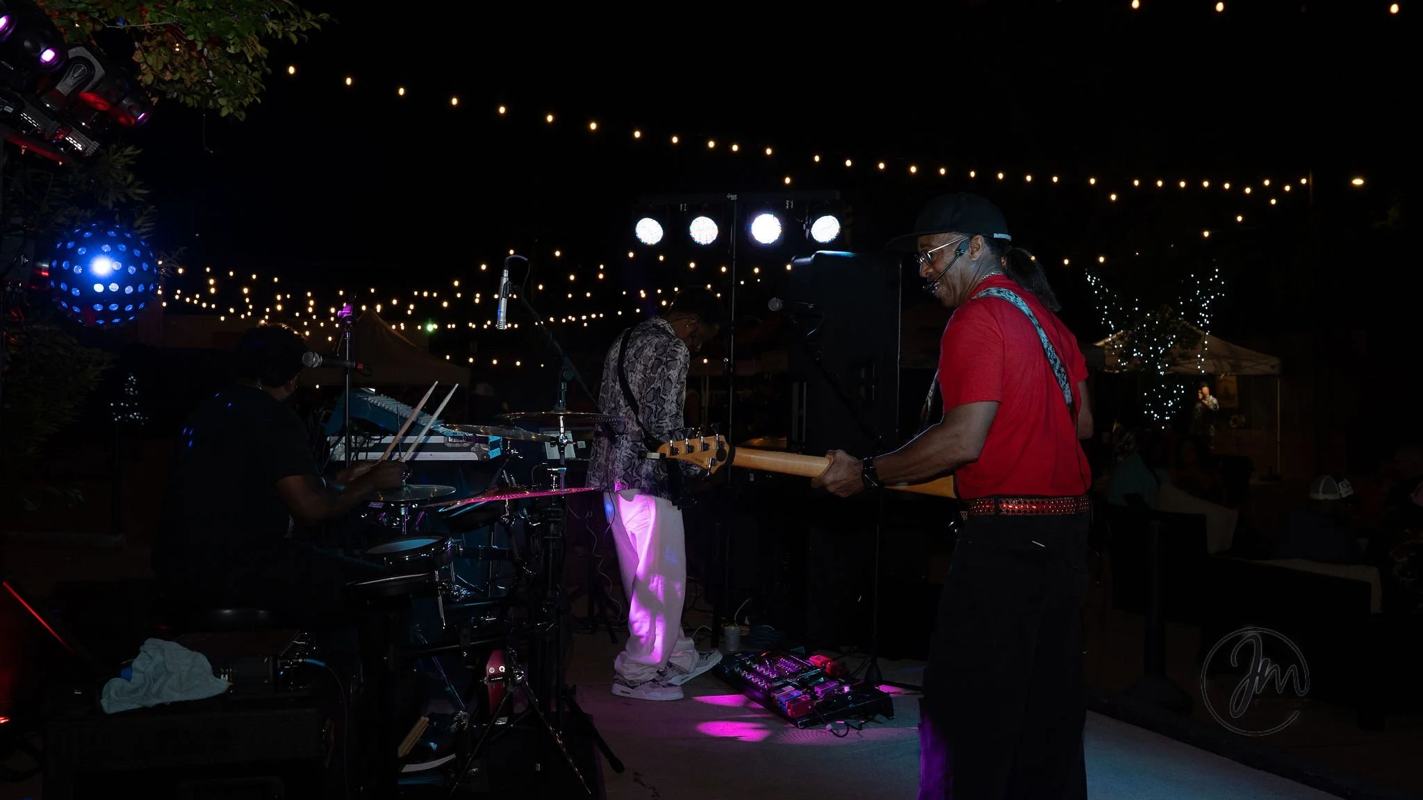 Band performing live outdoors at night under string lights, with three musicians playing instruments, including drums, bass, and guitar.