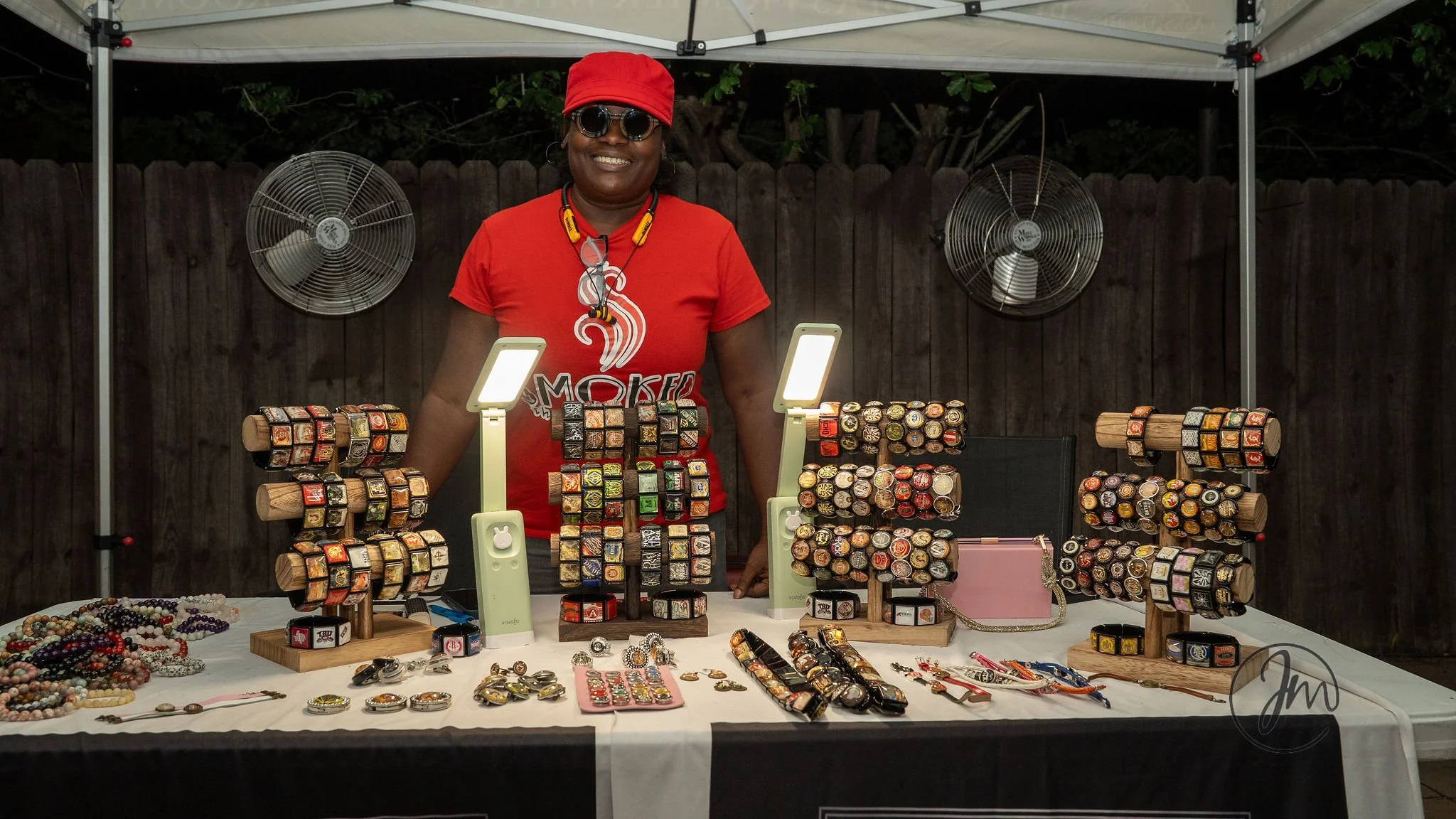 A woman in a red shirt and hat, wearing sunglasses, stands behind a table at an outdoor jewelry stall. The table displays various jewelry items including rings, bracelets, and necklaces, with some jewelry arranged on wooden display stands. The backgr