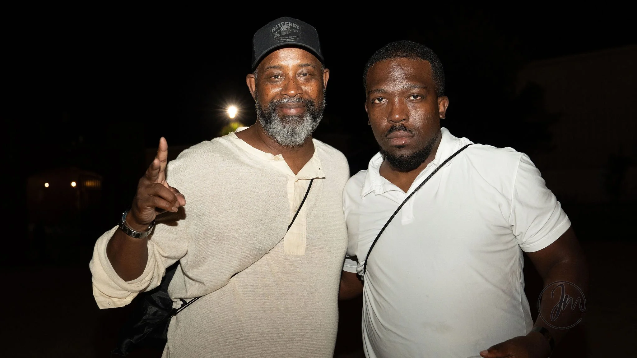 Two men standing close together at night, one with a beard wearing a hat and a light-colored shirt, making a peace sign, and the other with a serious expression wearing a white shirt.