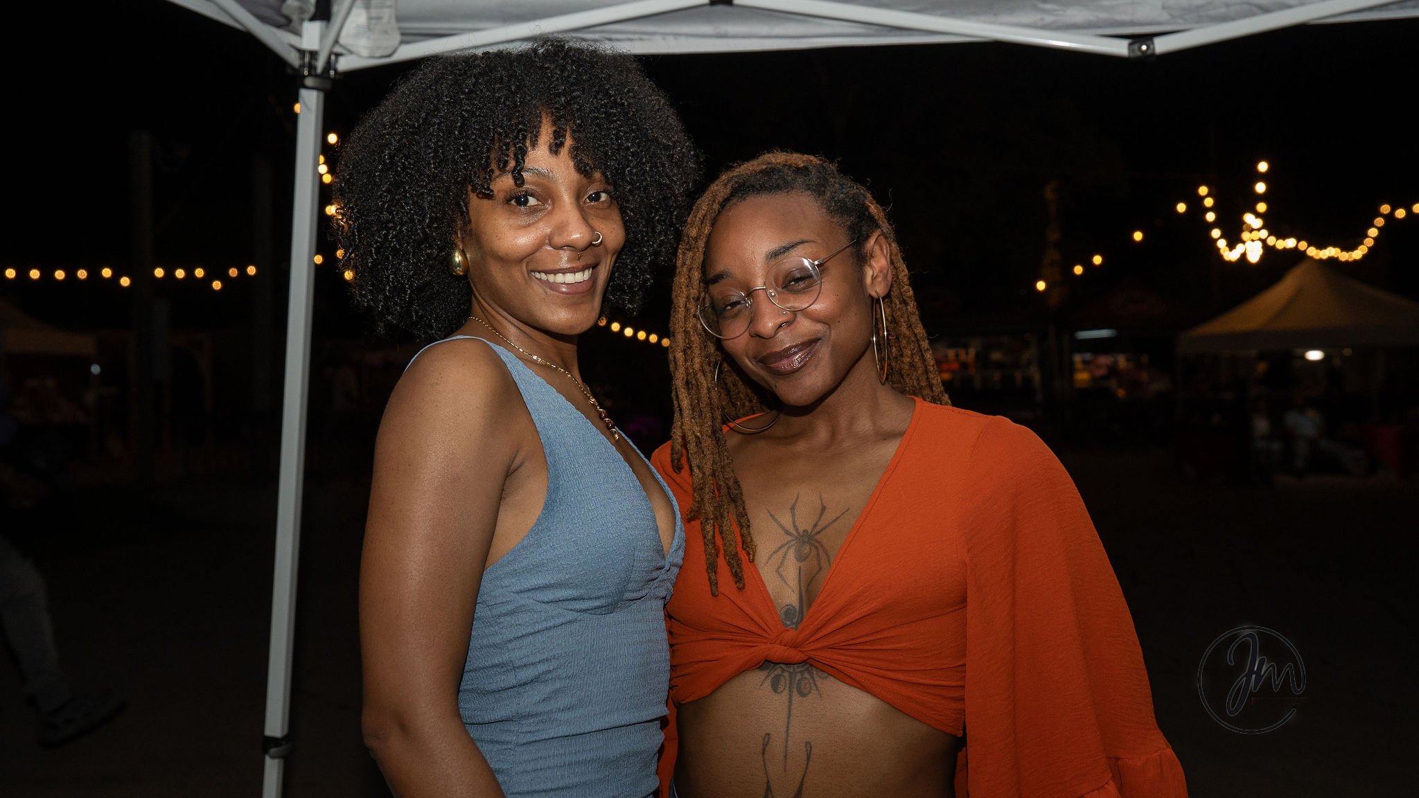 Two women standing close together at night during an outdoor event, smiling. The woman on the left has curly black hair, wearing a light blue sleeveless top. The woman on the right has dreadlocks, glasses, dark lipstick, an orange top with a tie-fron
