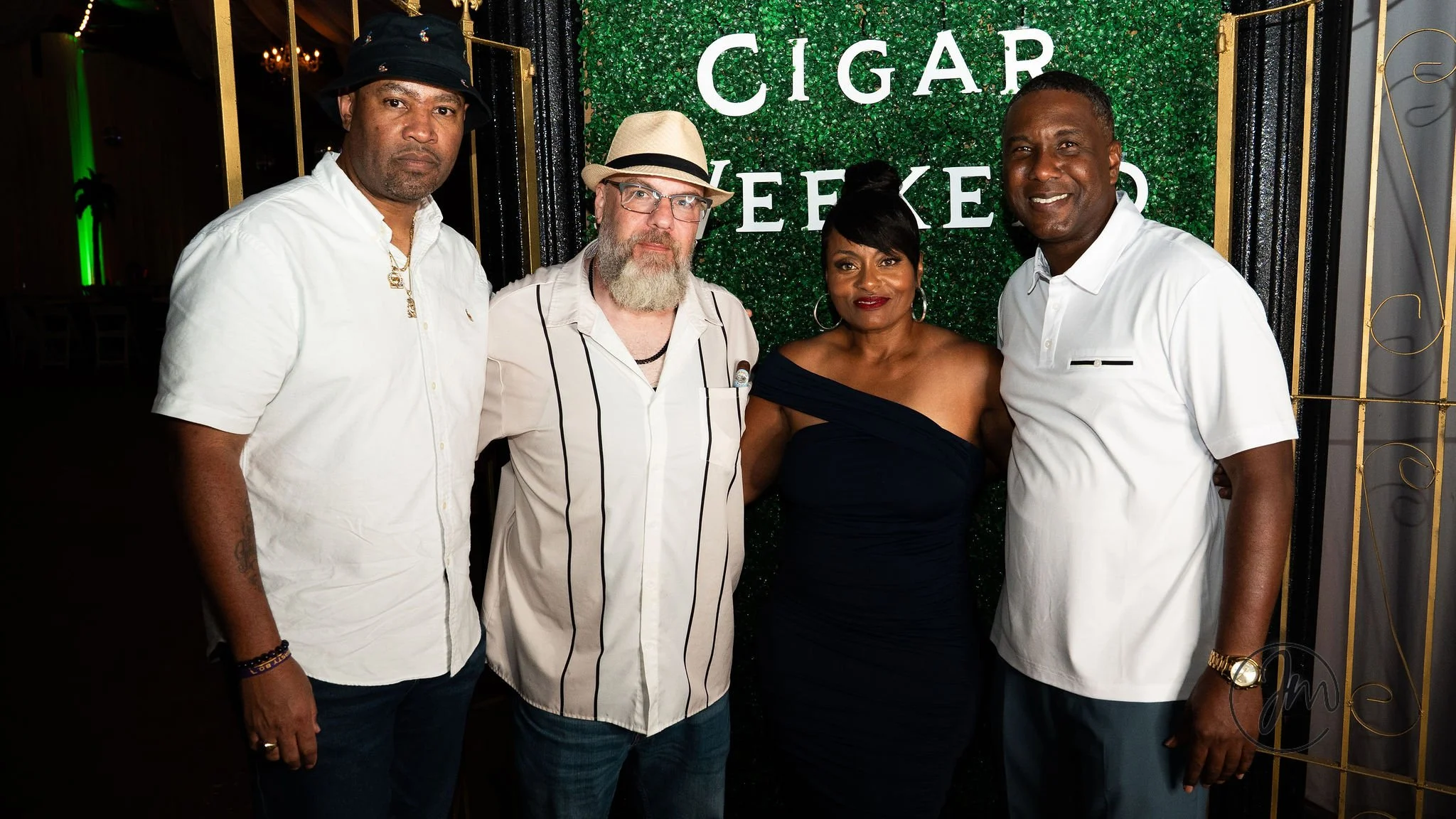 Four people standing in front of a green wall with the words 'CIGAR WEEKEND' behind them, smiling at the camera.