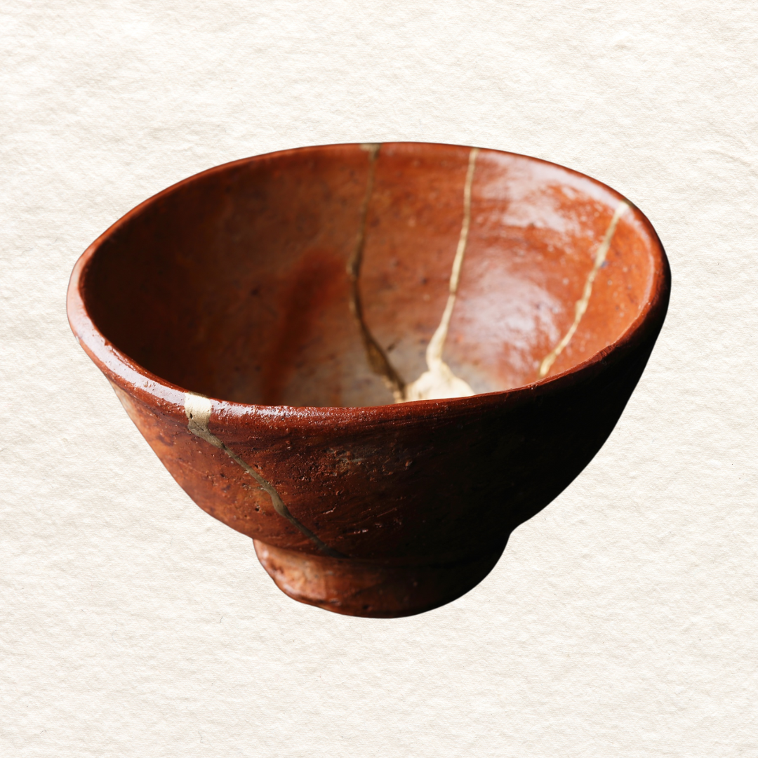 An ancient reddish-brown ceramic kintsugi bowl with visible cracks and gold inlay markings on its surface, placed against a plain background.
