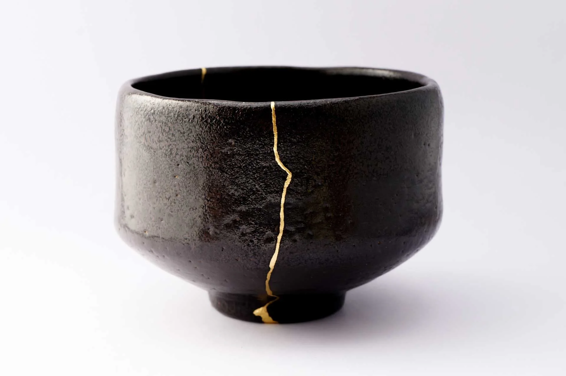 A black ceramic kintsugi bowl with a golden crack running from the top edge to the bottom edge, against a white background.