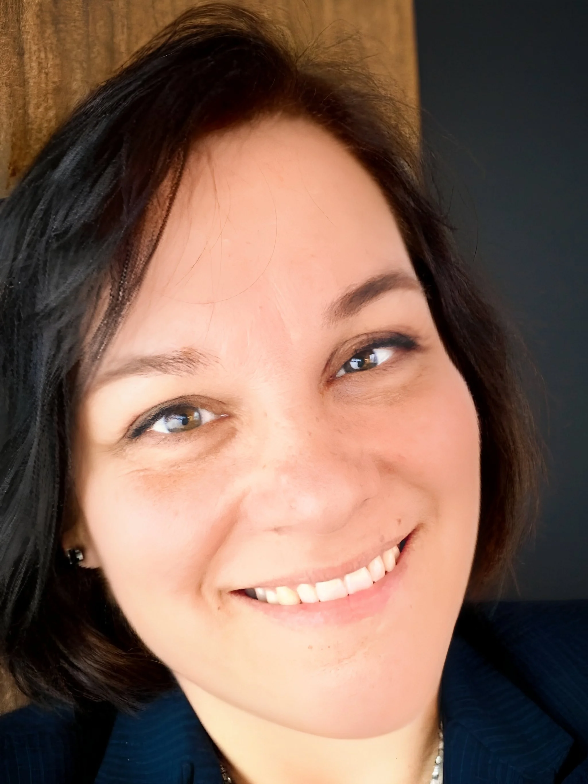 Close-up of a smiling middle-aged woman with dark hair, brown eyes, and light skin, wearing a blue top and a pearl necklace.