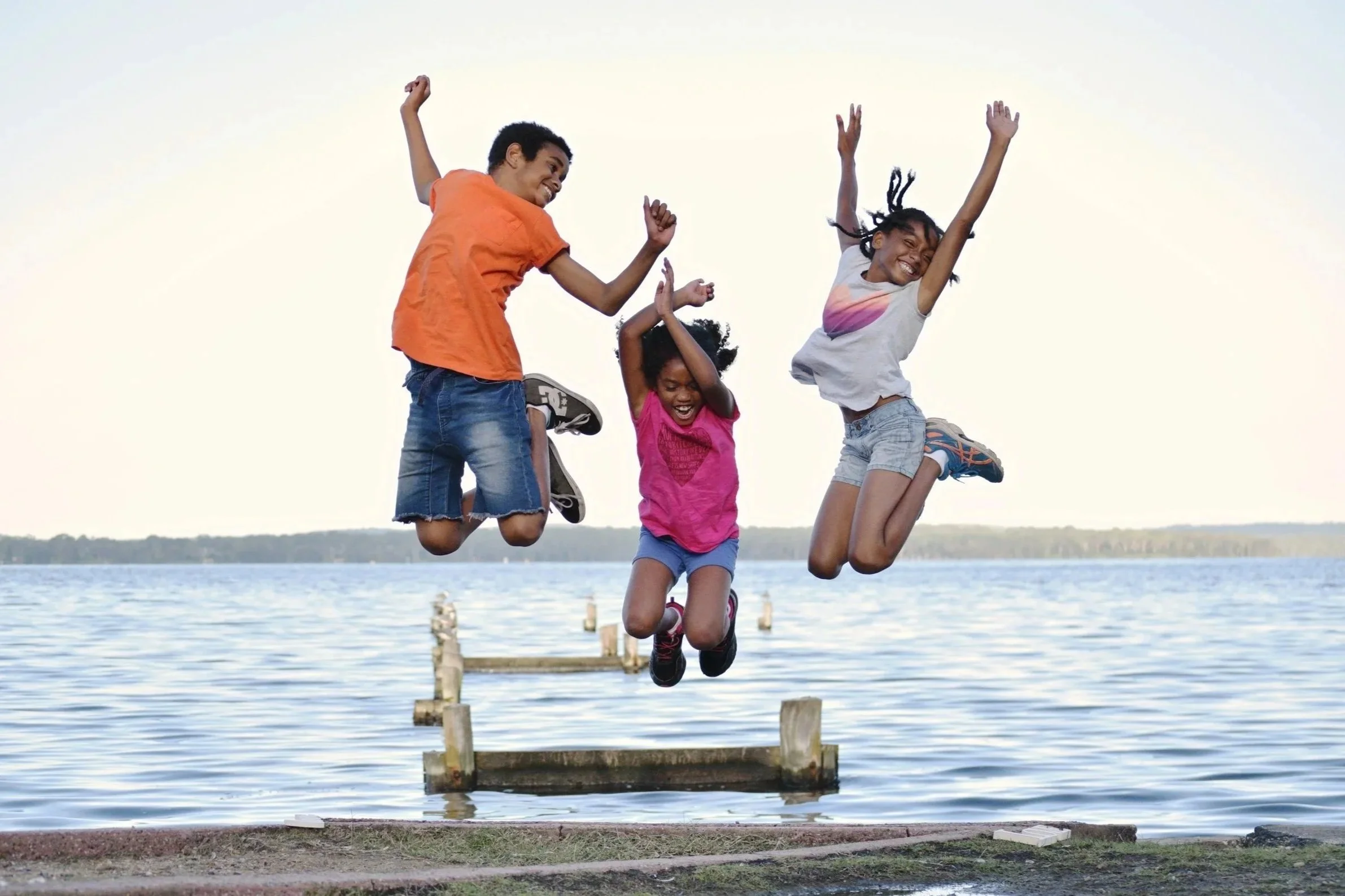 Three children doing yoga in a tree pose, standing on one leg with hands together overhead, against a white wall and green floor.