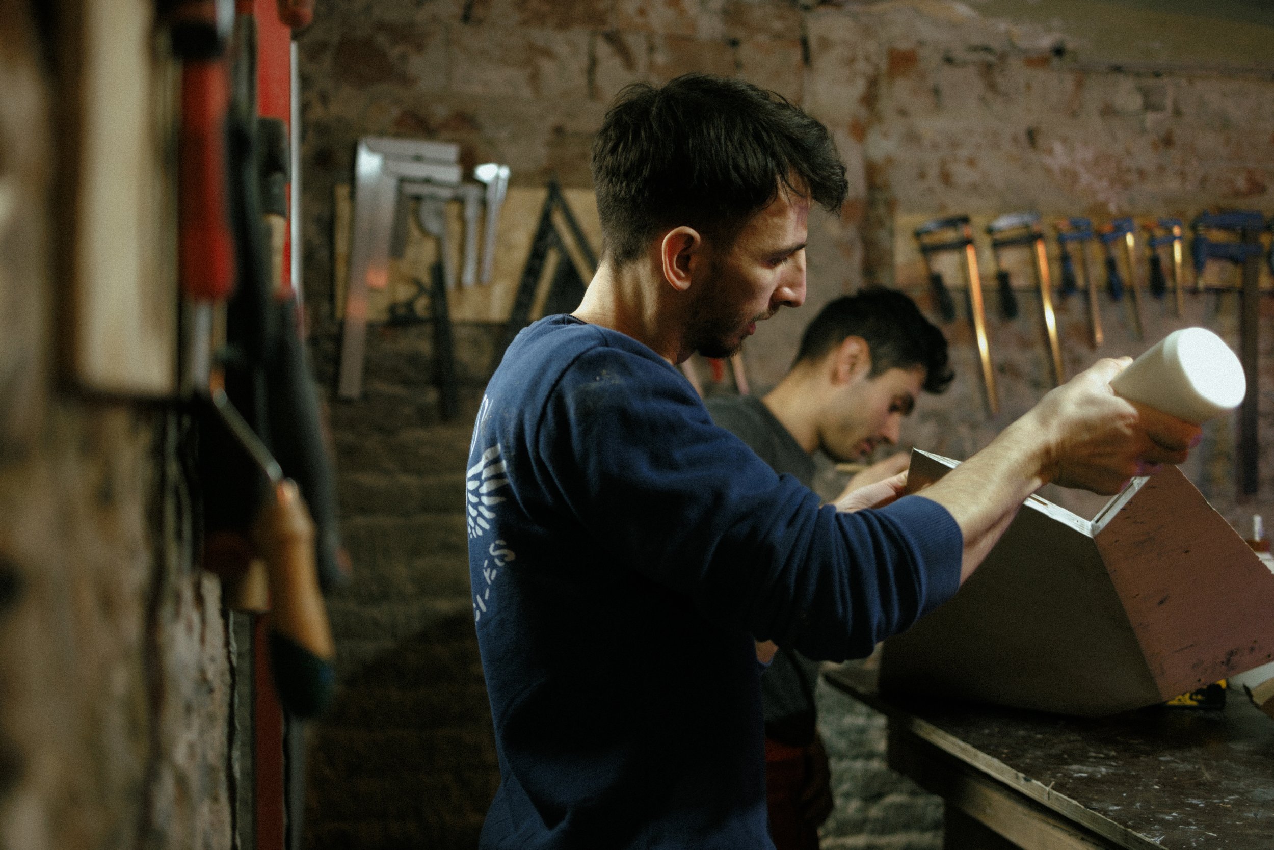 Two men working on a woodworking project in a workshop, one applying glue to a wooden piece; tools visible in the background.